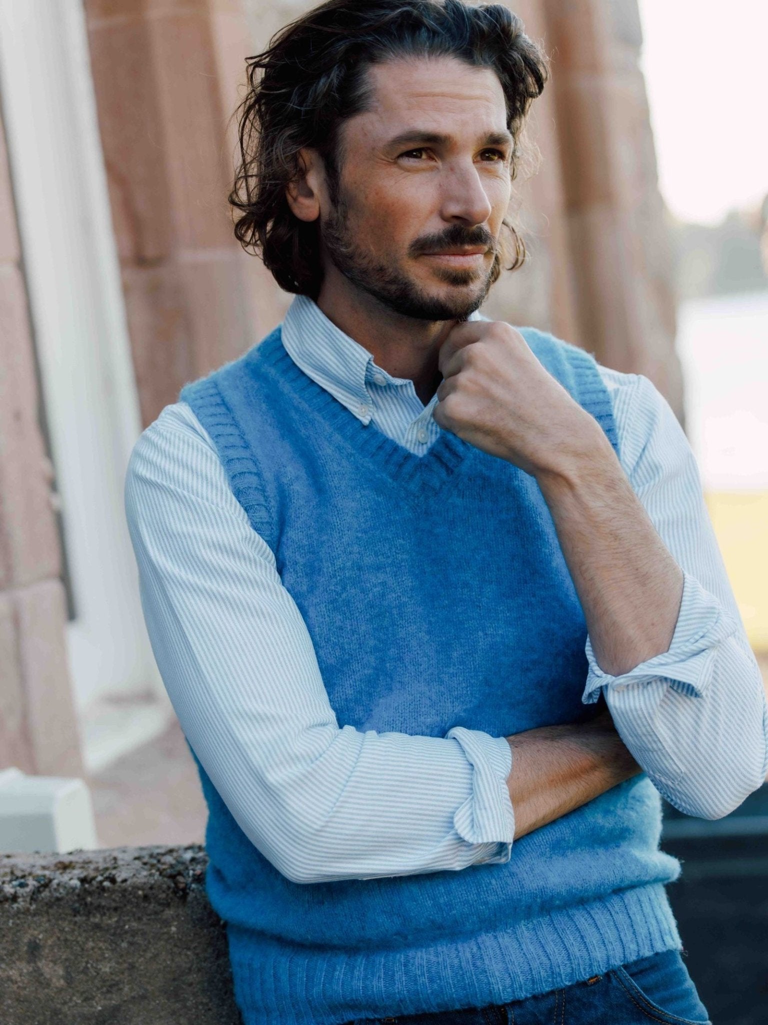 A man with wavy brown hair and a trimmed beard stands outdoors, resting one arm on a ledge and touching his chin. He wears a Campbells of Beauly Oxford Shirt under a light blue sweater vest, gazing thoughtfully into the distance.