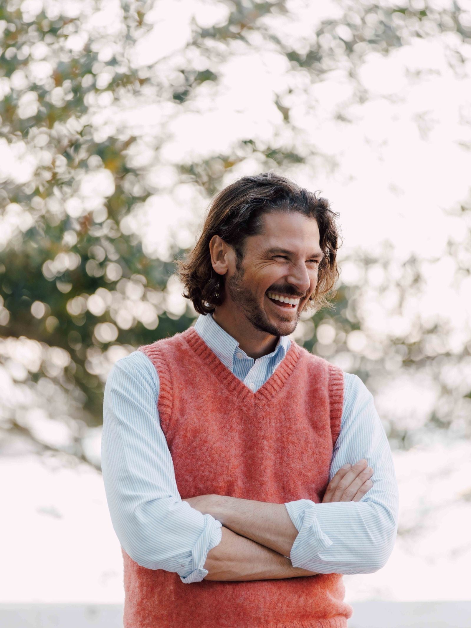 A bearded man with shoulder-length hair stands outdoors, smiling in a light blue shirt and a coral Shetland Slipover by Campbells of Beauly, a classic knit vest. The bright background is blurred with lush greenery.