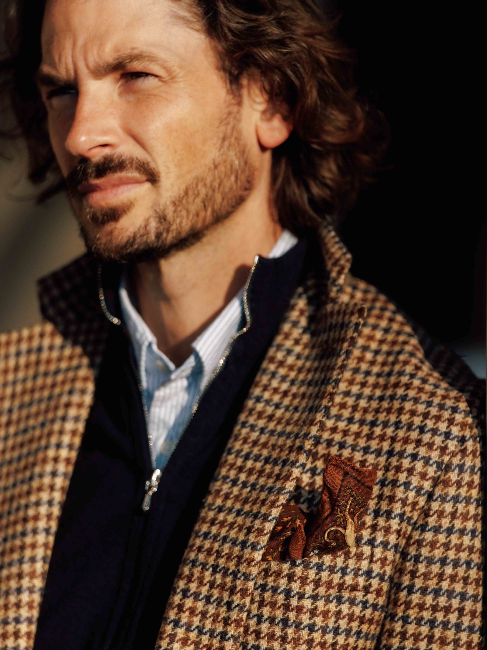 A man with wavy brown hair and a beard wears a Campbells of Beauly Navy Gun Club Tweed Jacket, paired with a light blue shirt, dark zip-up sweater, and patterned brown pocket square.