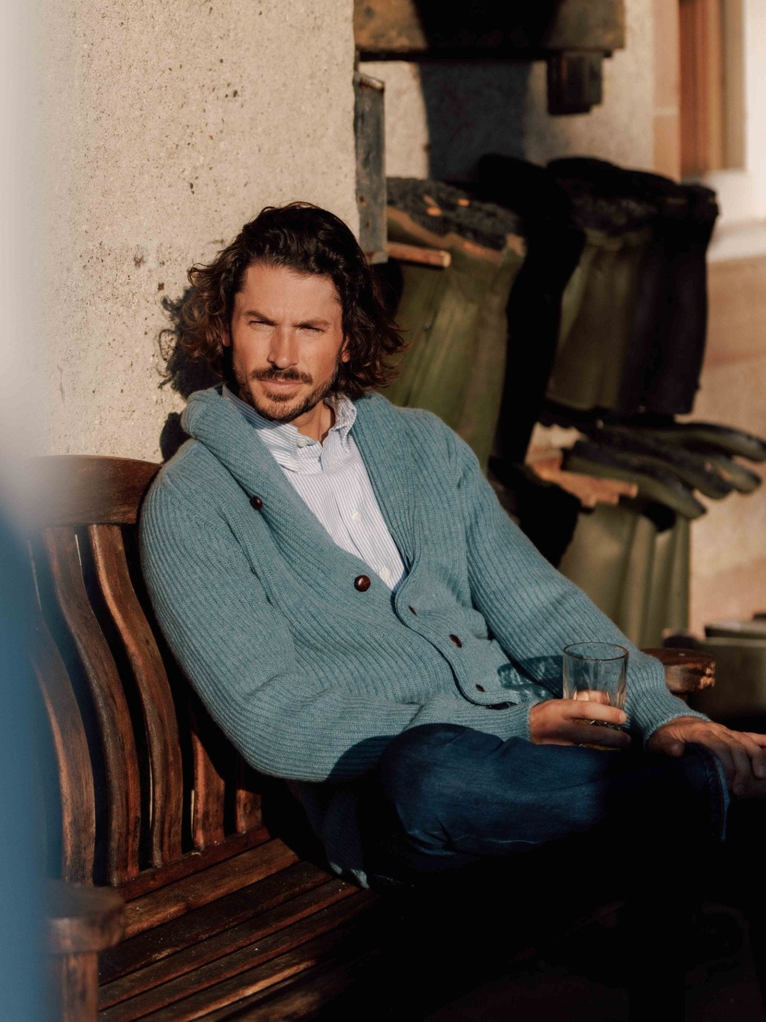 A man with curly hair sits on a wooden bench, wearing Campbells of Beauly Shawl Collar Cardigan in light blue over a collared shirt and jeans. Sunlight casts shadows behind him onto the wall and garden tools.
