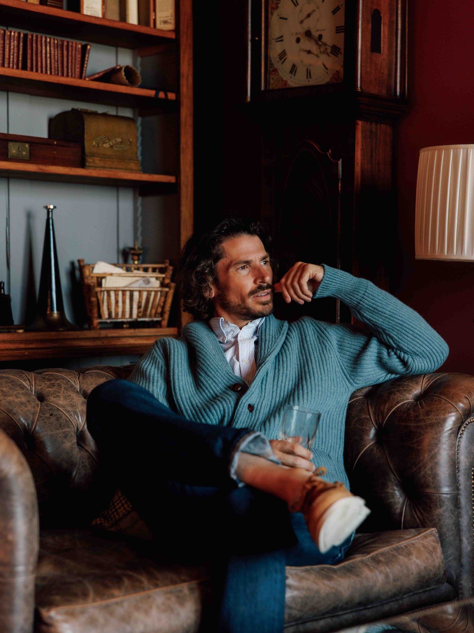 A man with curly hair wears the Campbells of Beauly Shawl Collar Cardigan over a white shirt and jeans as he sits on a brown leather sofa holding a glass, with shelves, books, baskets, a large clock, and a tall lamp in the background.