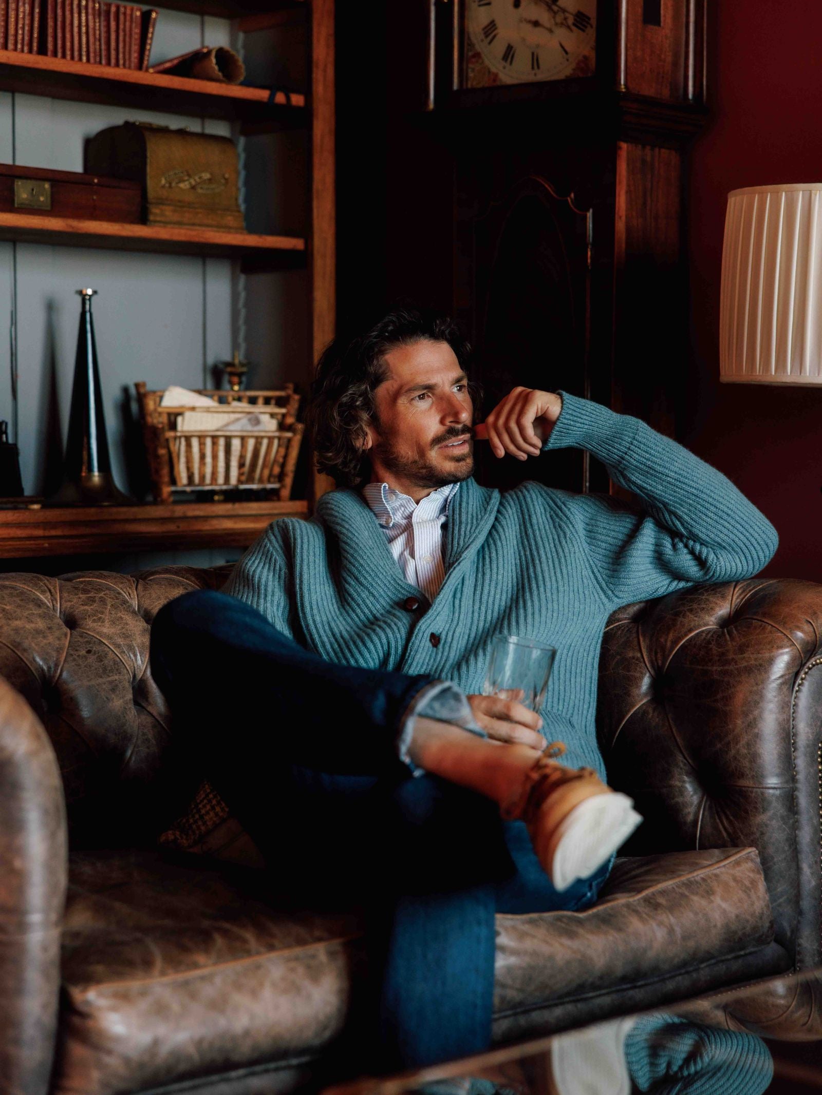 A man with wavy hair, wearing a light blue Campbells of Beauly Cashmere Shawl Neck Collar Cardigan, shirt, and jeans, sits on a brown leather sofa with a glass. Behind him are wooden shelves holding books, a clock, and decor.