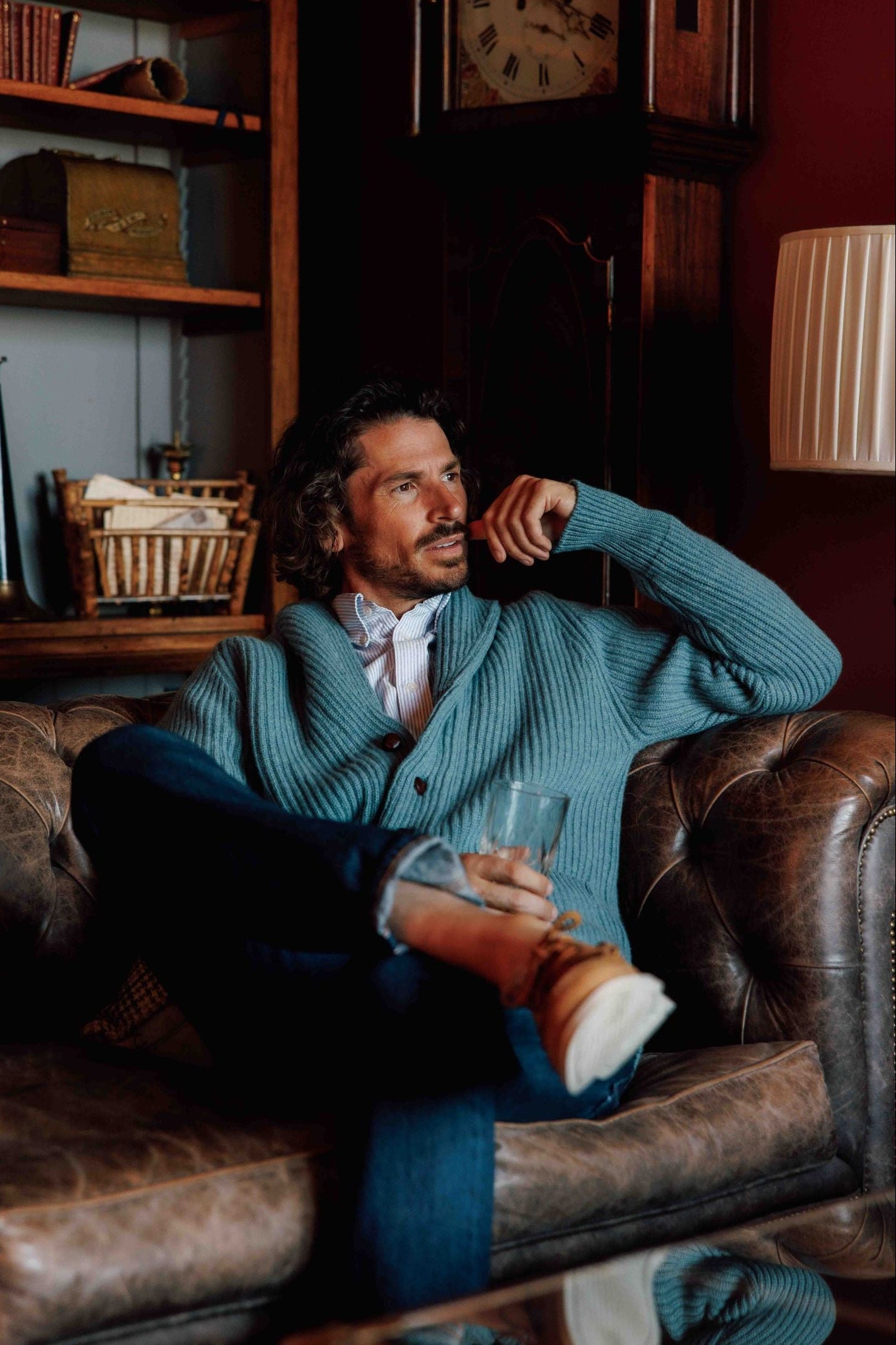 A man with wavy hair, wearing a light blue Campbells of Beauly Cashmere Shawl Neck Collar Cardigan, shirt, and jeans, sits on a brown leather sofa with a glass. Behind him are wooden shelves holding books, a clock, and decor.