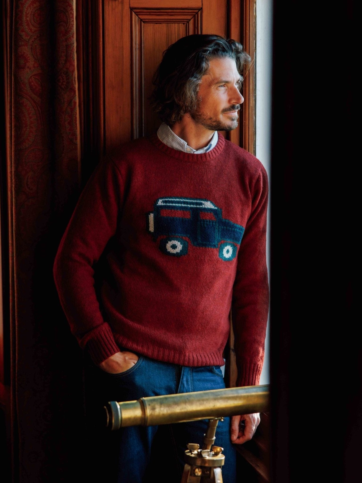 A man with wavy hair and a beard gazes out of a window, wearing the Campbells of Beauly Land Rover Defender Jumper—a classic red sweater with a blue car motif—standing beside a brass telescope in a wood-paneled room.