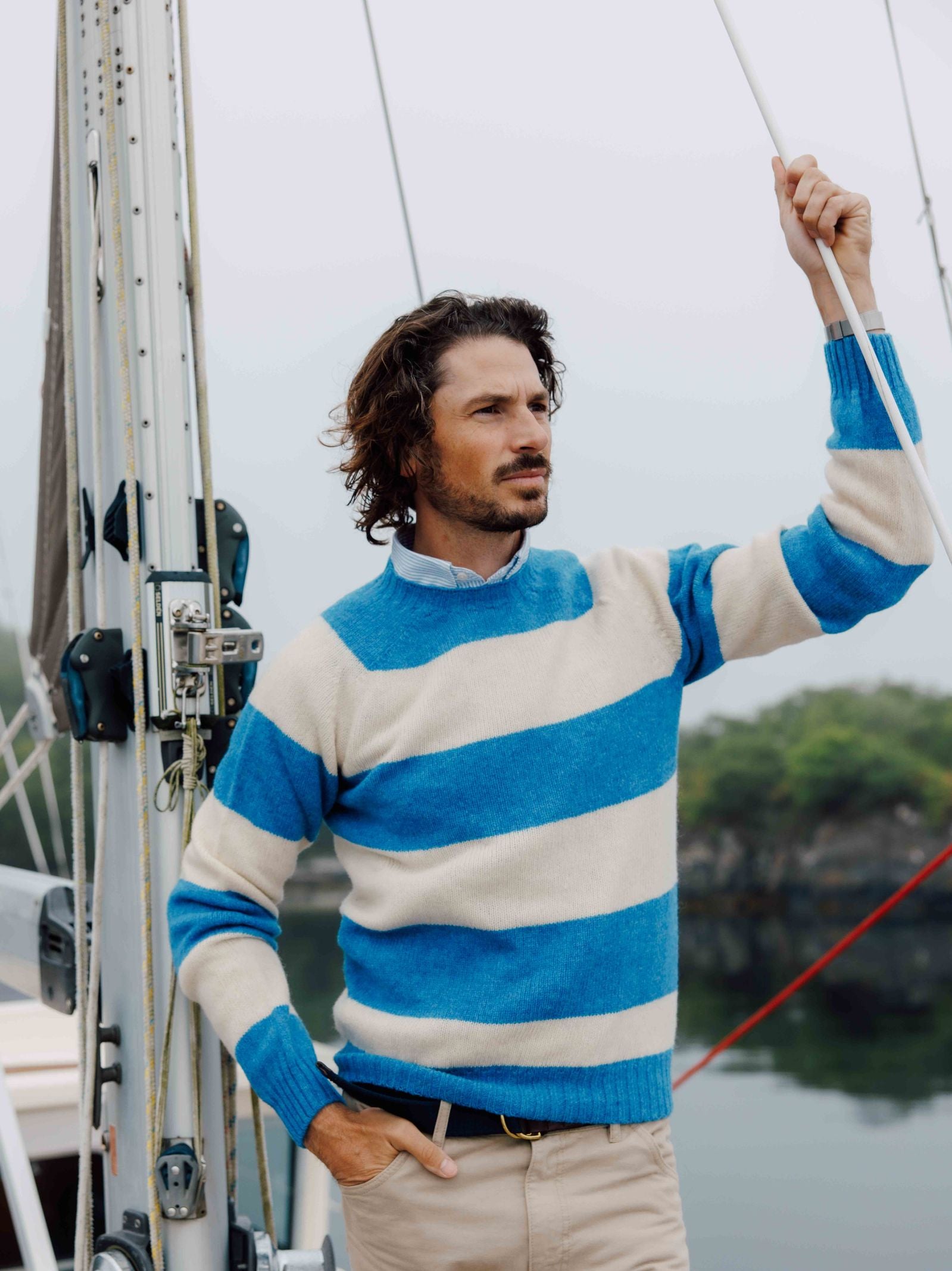 A man with wavy hair wears the Campbells of Beauly Shetland Stripe Crew Jumper in blue and white as he stands on a boat, holding a rope and gazing into the misty distance, surrounded by trees and calm water.