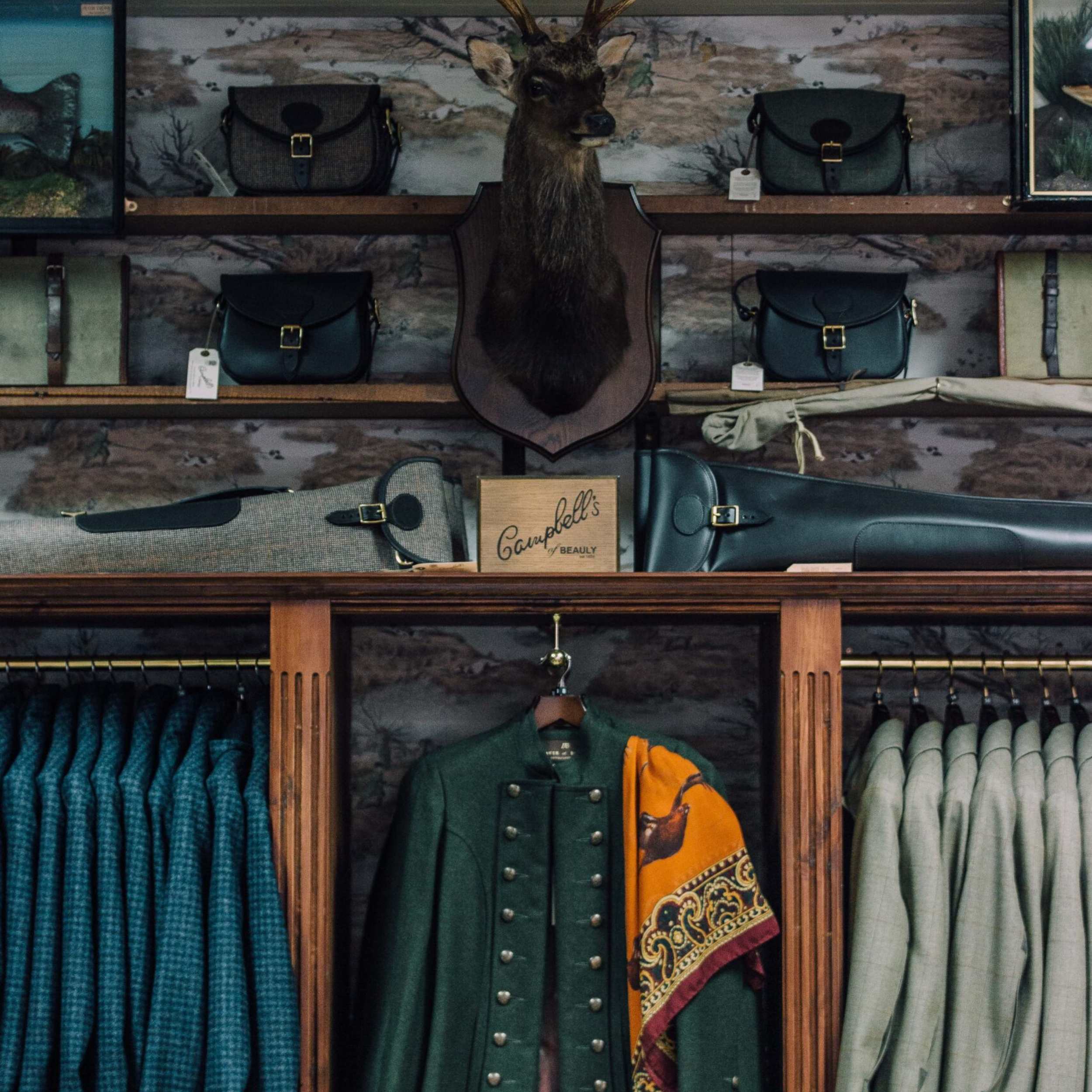 A clothing store display featuring plaid jackets, a green coat, a yellow scarf, and various handbags is set against wooden shelving with a mounted deer head and the signature Campbell’s of Beauly sign.