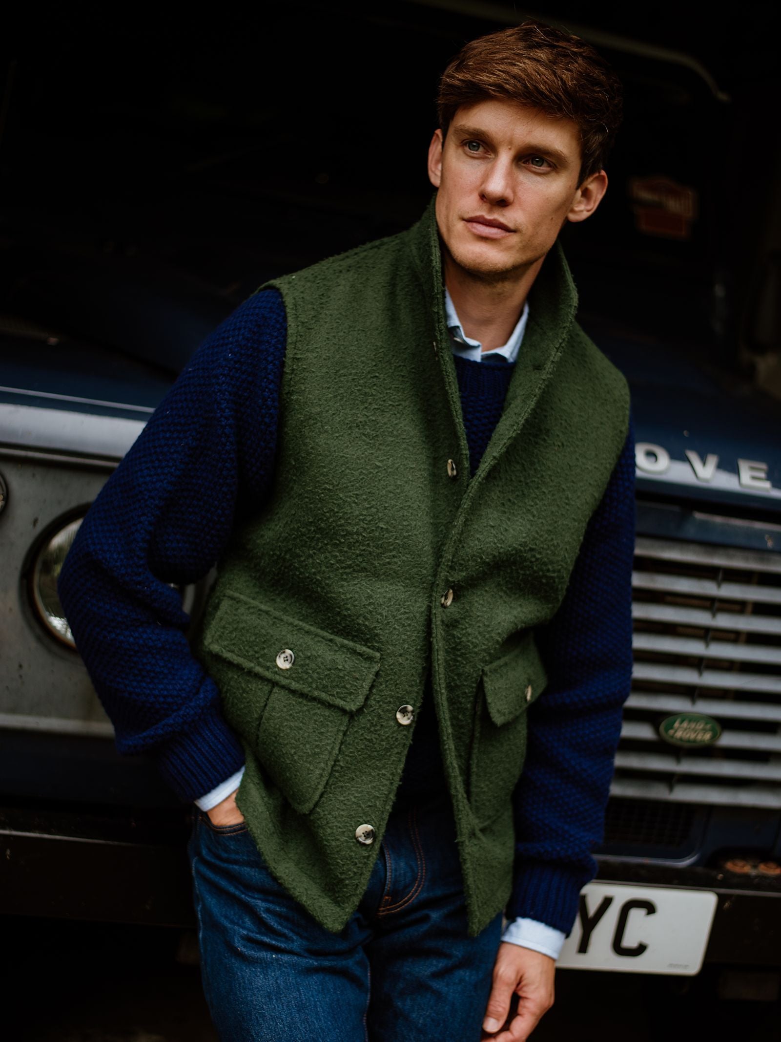 A man stands in front of a vintage blue Land Rover, wearing the Campbell’s of Beauly Casentino Fleece Gilet over a blue sweater and jeans, with his hand in his pocket, looking thoughtfully to the side.