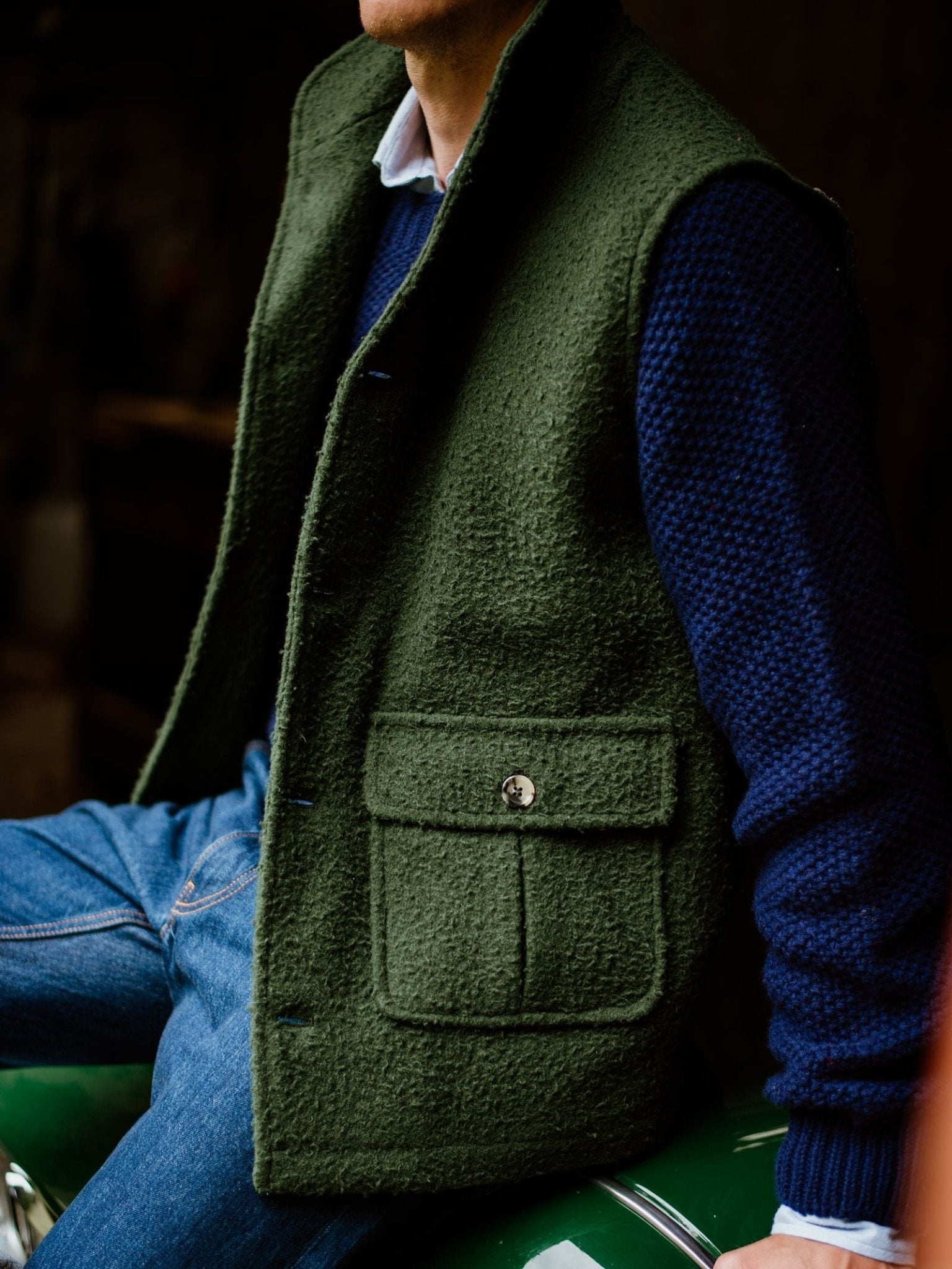 A person wearing the Campbell’s of Beauly Casentino Fleece Gilet in textured green, layered over a navy blue sweater and blue jeans, sits on a green surface. Only the lower face and body are visible against a dark, out-of-focus background.
