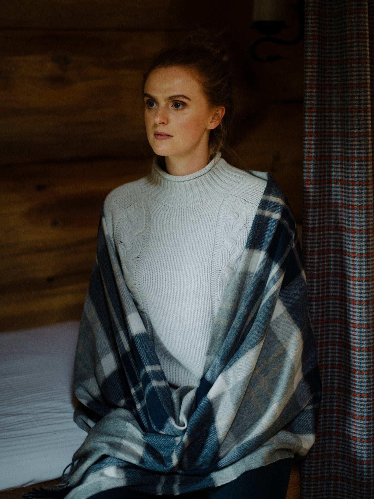 A woman with light brown hair sits indoors on a bed, wrapped in a Campbells of Beauly Cashmere Stole. She gazes calmly to the side, with wooden walls and a plaid curtain evoking the charm of the Scottish Borders.
