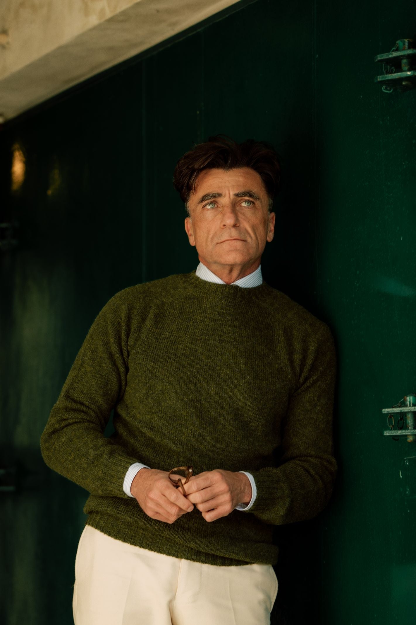 A man with dark hair stands against a dark green door, wearing Campbells of Beauly Shetland Jumper in green over a collared shirt and cream pants, looking slightly upward while holding eyeglasses.