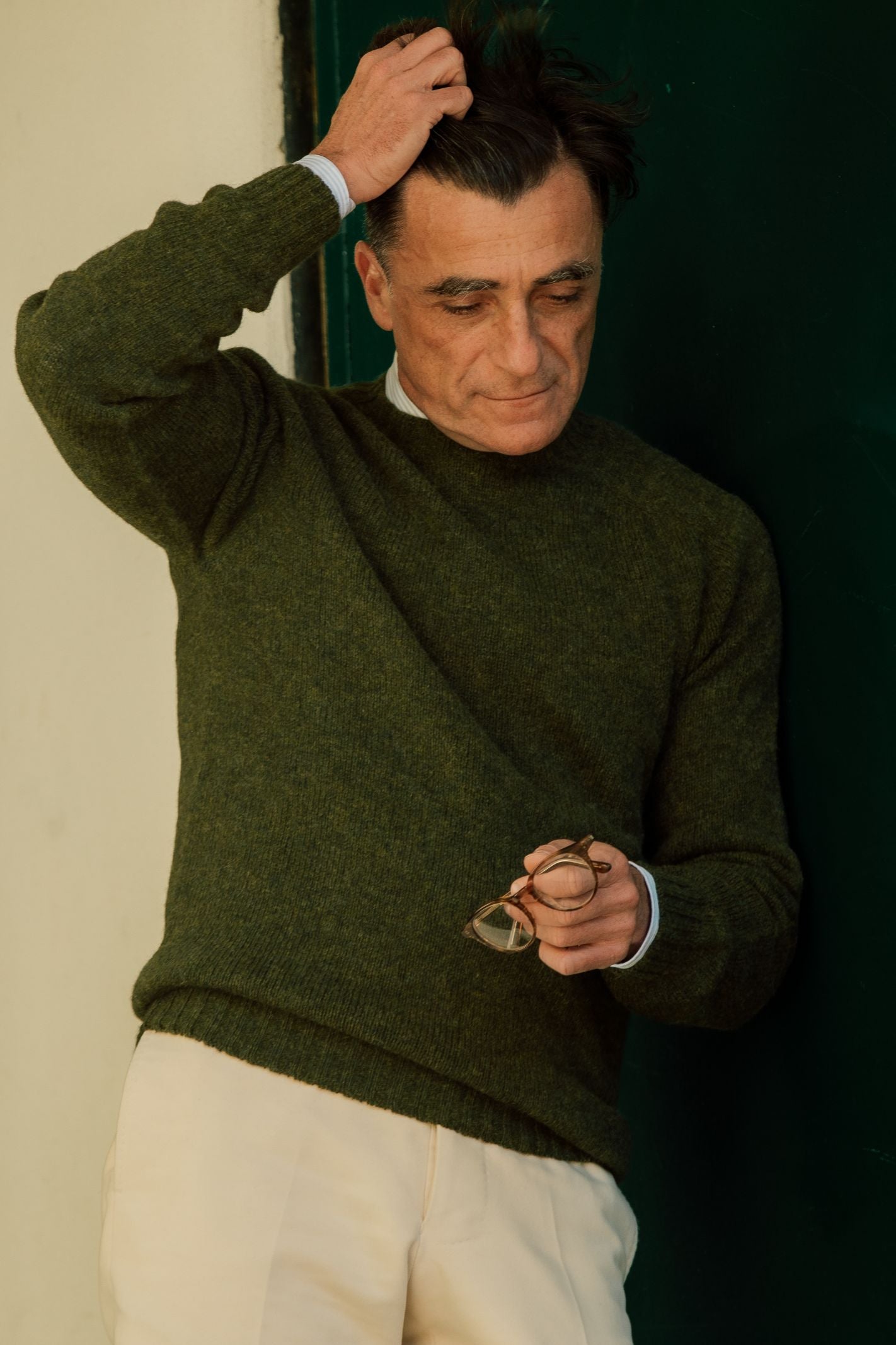 A middle-aged man in a Campbells of Beauly Shetland Jumper in green stands against a dark wall, holding glasses in one hand and running his other hand through his hair, looking down with a soft smile.
