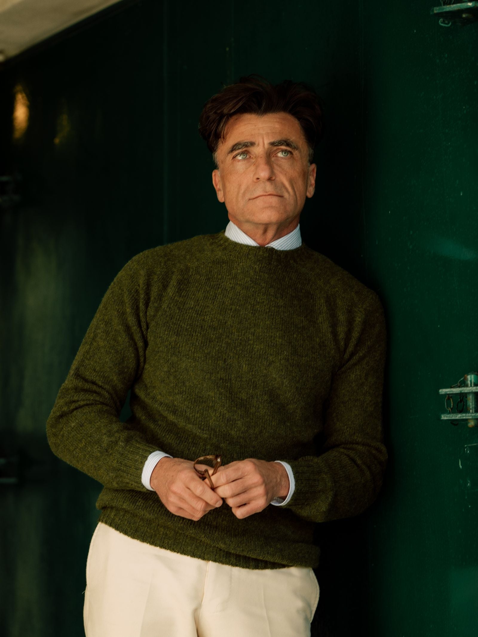 A middle-aged man with brown hair wears a green Campbell's of Beauly Shetland Jumper over a collared shirt and white pants, standing against a dark green wall, holding eyeglasses and gazing upward with a calm expression.