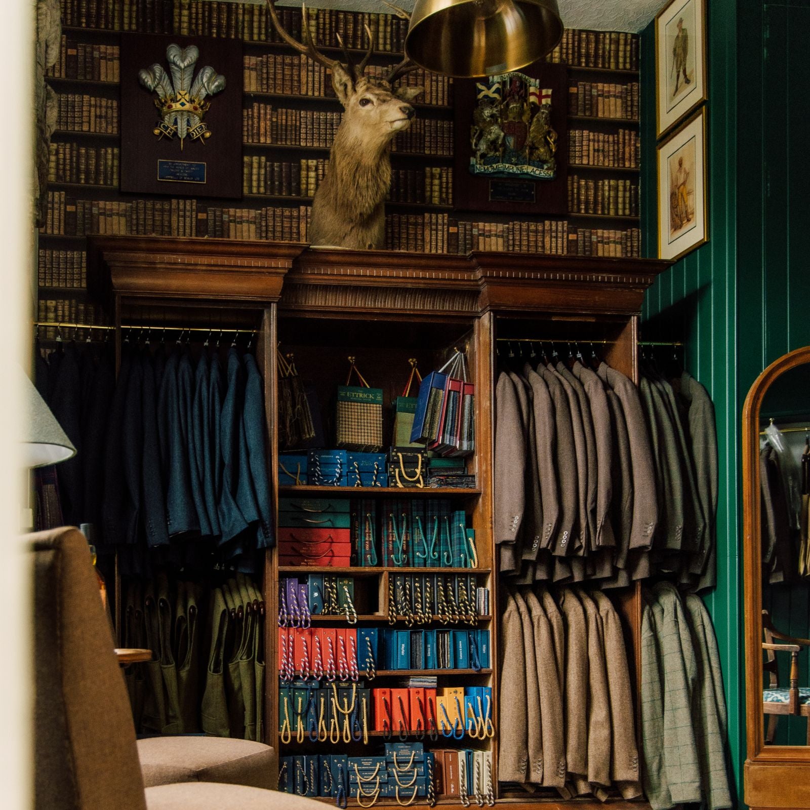 A vintage clothing shop with neatly hung suits, colorful ties, and belts, wooden shelves, and a mounted deer head above. The walls are decorated with patterned wallpaper and framed art, creating a classic, elegant atmosphere.
