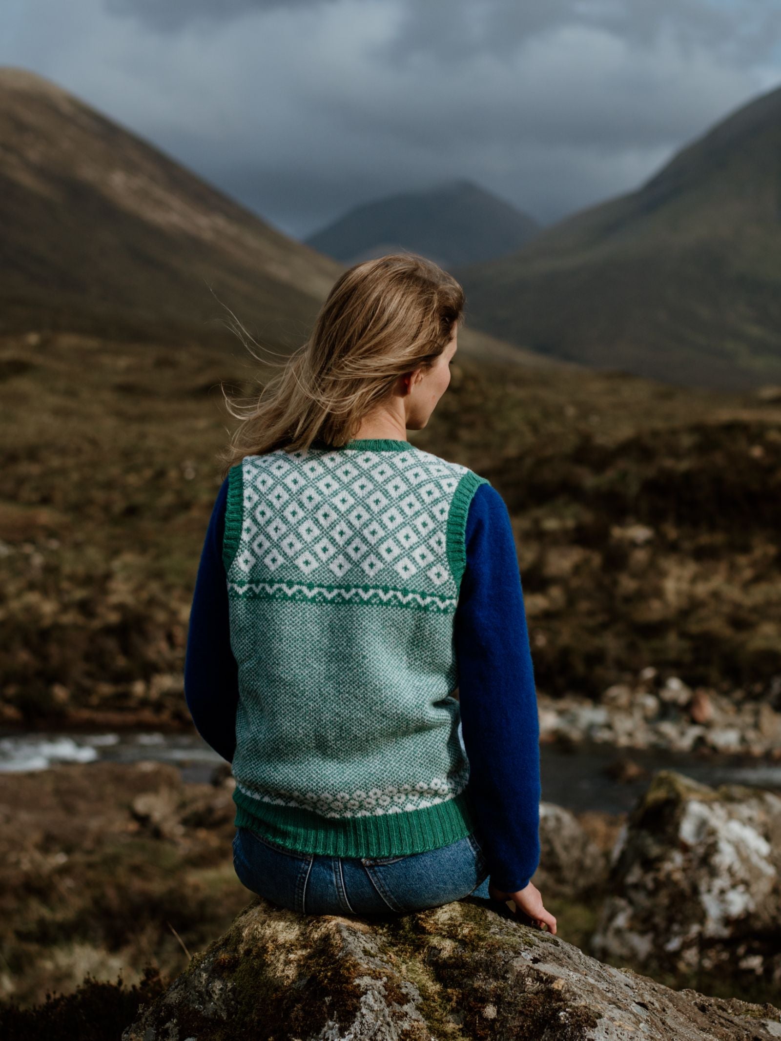 A person with long blonde hair, wearing Campbells of Beaulys Nordic V Tank, sits on a rock facing away, gazing over a river and lush mountains under a cloudy sky.