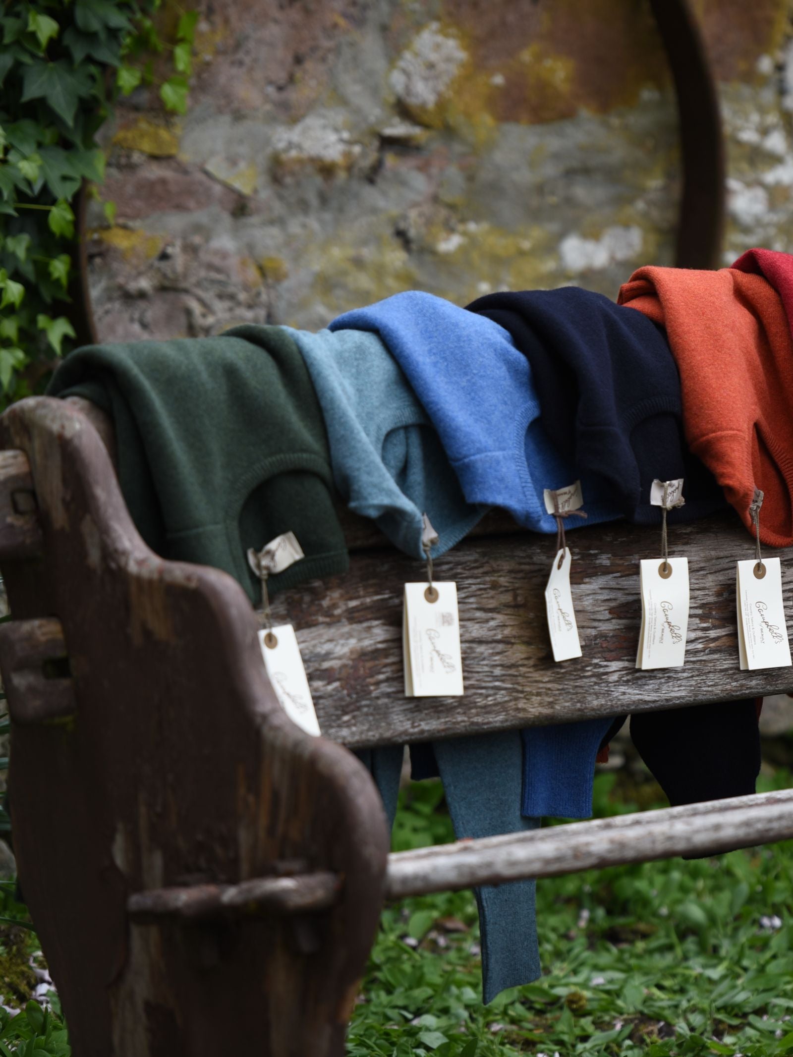 Five folded Campbell’s of Beauly Lambswool Crew Neck Jumpers in green, gray, blue, black, and orange lie on a rustic wooden bench outdoors, each with a white tag and a mossy Scottish Borders stone wall behind.
