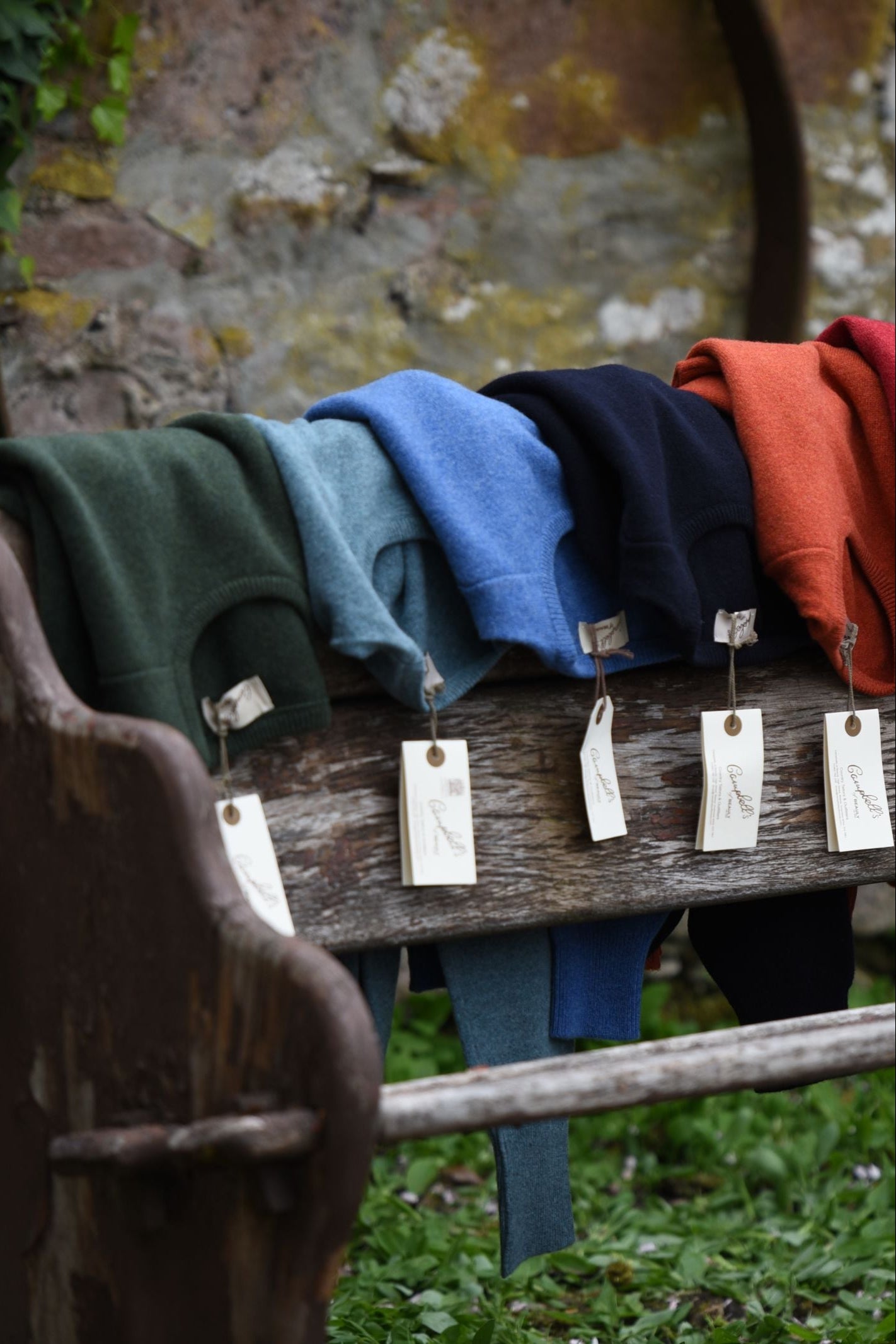 Five folded Campbells of Beauly Lambswool Crew Neck Jumpers in green, teal, blue, navy, and orange—each with a tag attached—are displayed on a rustic wooden bench outdoors in the Scottish Borders.