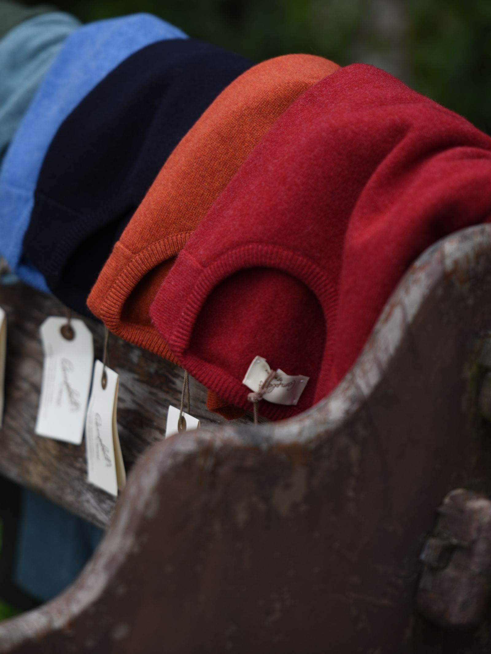 A row of Campbells of Beauly Lambswool Crew Neck Jumpers in blue, black, orange, and red sits on a rustic wooden bench. Each has a tag attached, evoking cozy Scottish Borders style and suggesting they are for sale.