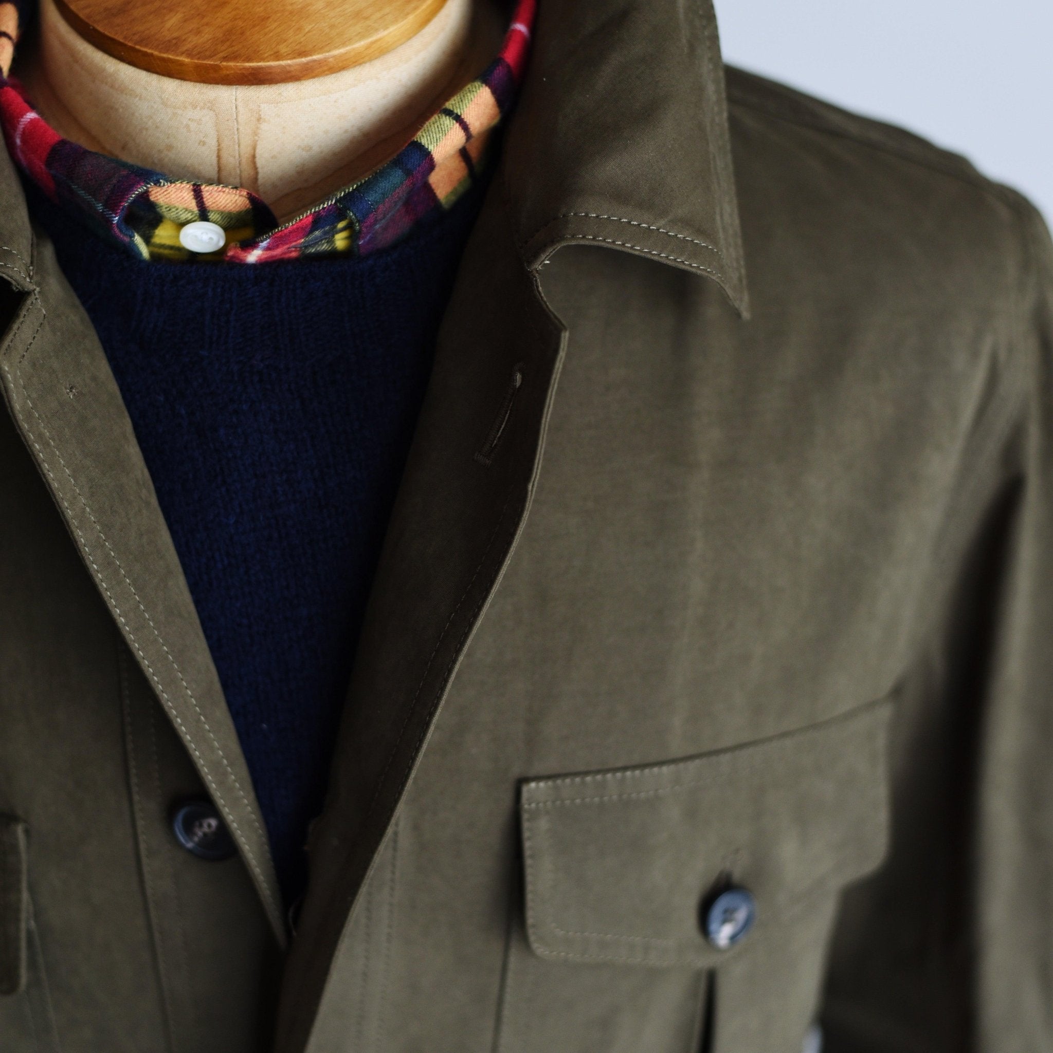 Close-up of a mannequin wearing Campbells of Beauly Aigas Field Jacket in olive green, featuring large buttons, front pockets, water-repellent finish, styled with a navy sweater and colorful patterned scarf.