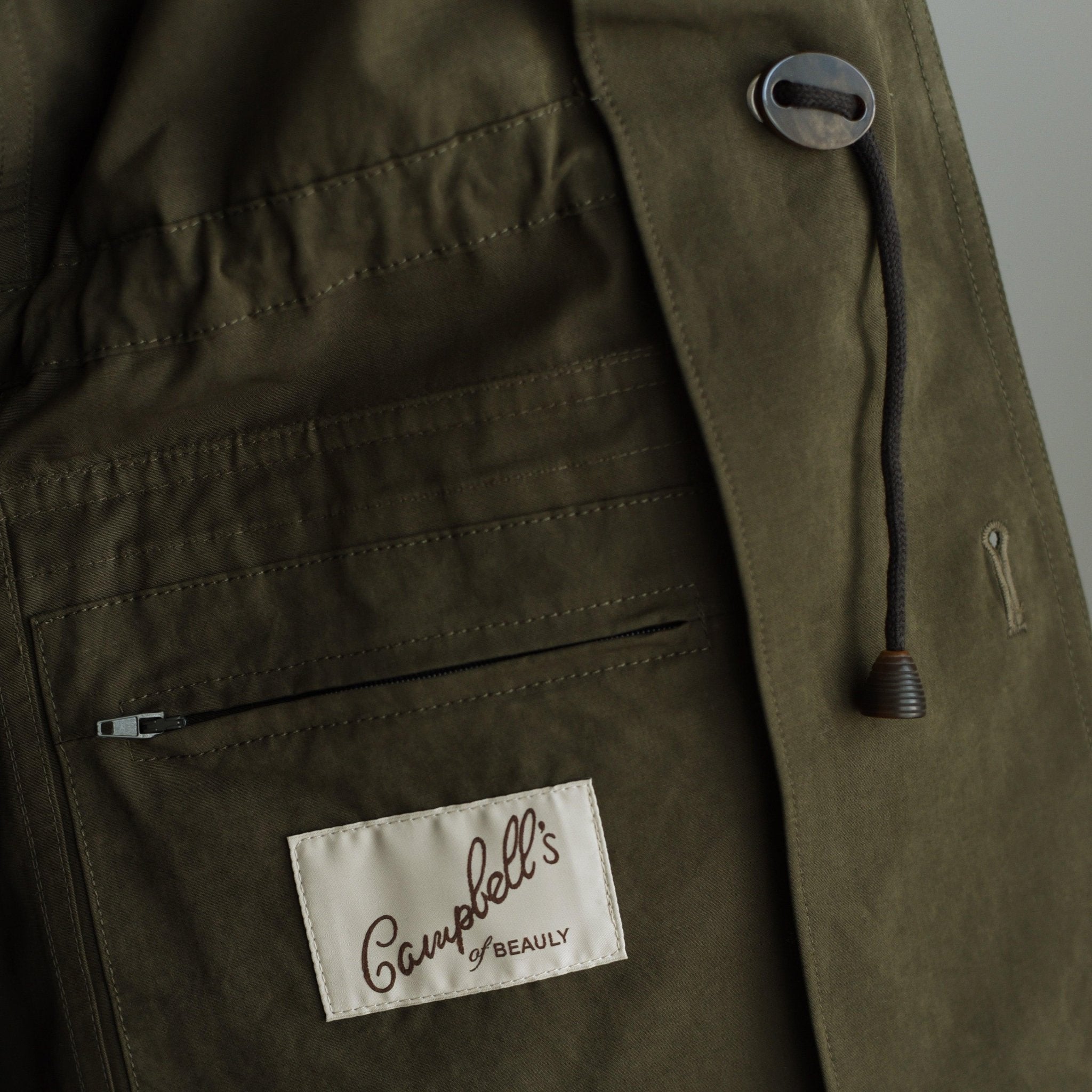 Close-up of the Campbells of Beauly Aigas Field Jacket in olive green, with a zippered pocket, sewn-on brand label, black drawstring with toggle, and made from soft peach skin fabric.