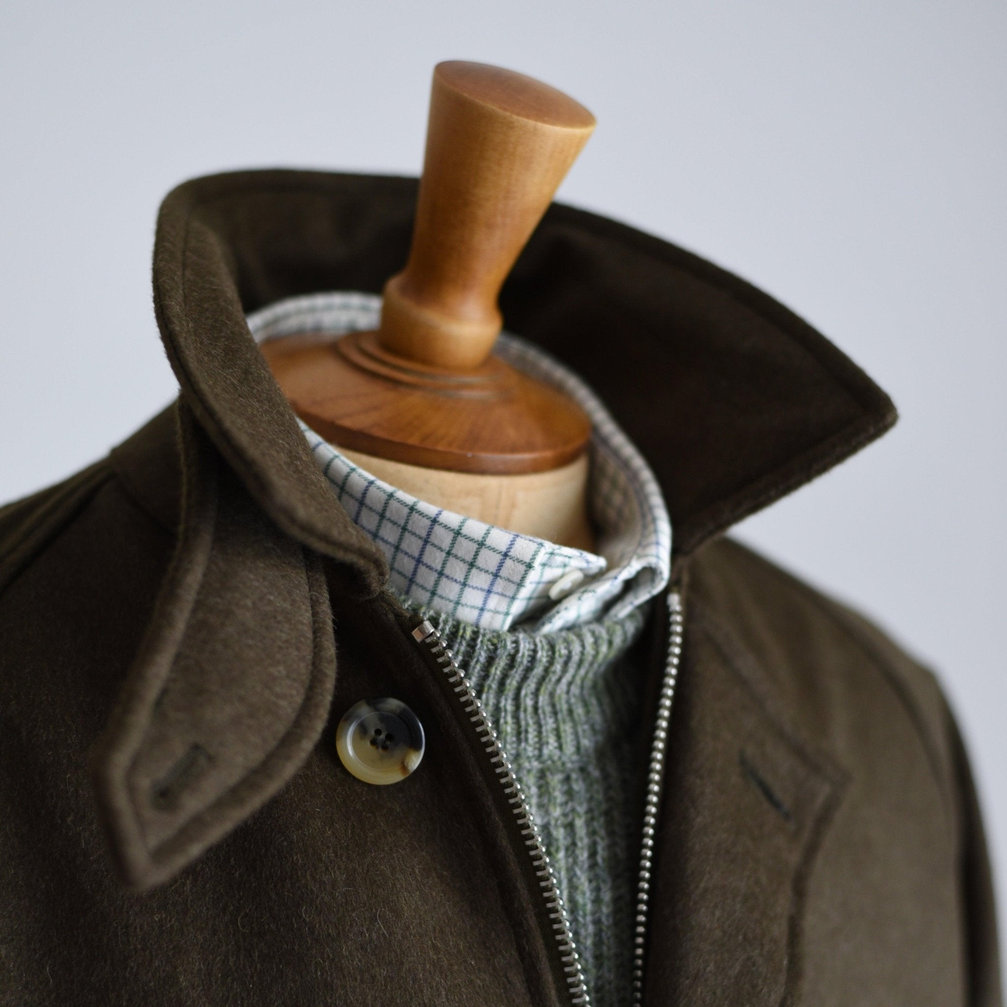 A close-up shows a mannequin in the Campbells of Beauly Cannich Coat, dark olive with a raised collar, layered over a green knit sweater and white checkered shirt. The mannequin’s wooden neck stands out against a plain, light background.