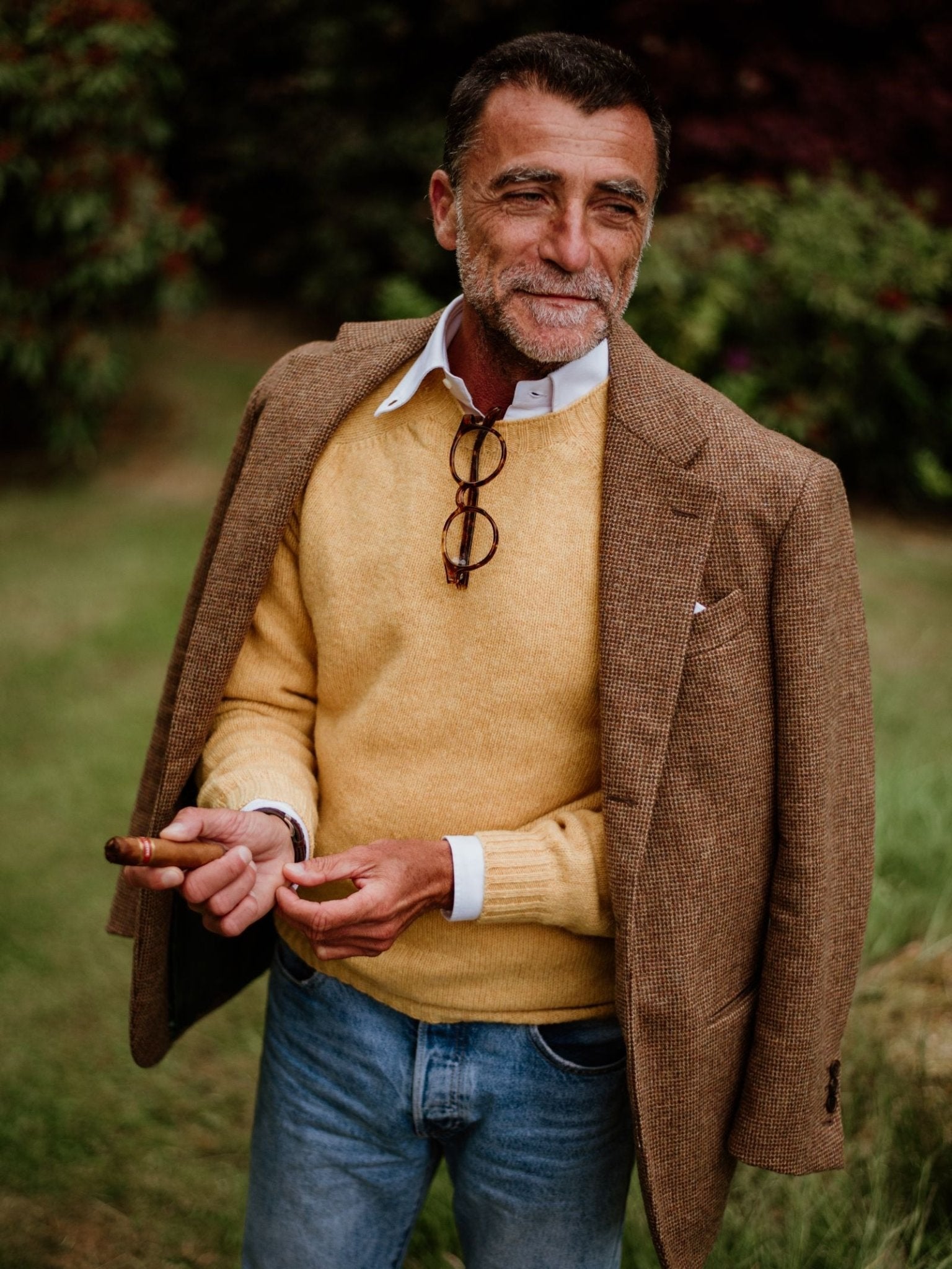 A middle-aged man with a beard stands on grass, smiling slightly as he wears Campbell's of Beauly Shetland Jumper in yellow under a brown blazer and white shirt. Glasses hang from his collar, and he holds a cigar; shrubs are visible behind him.