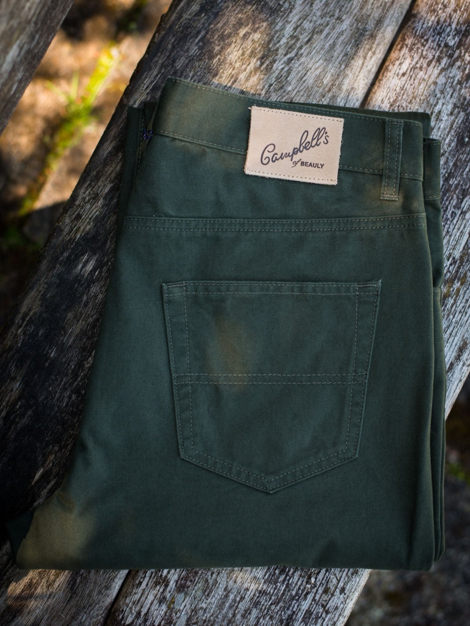 A pair of folded dark green Colour Cotton Jeans from Campbell's of Beauly rests on a weathered wooden surface.