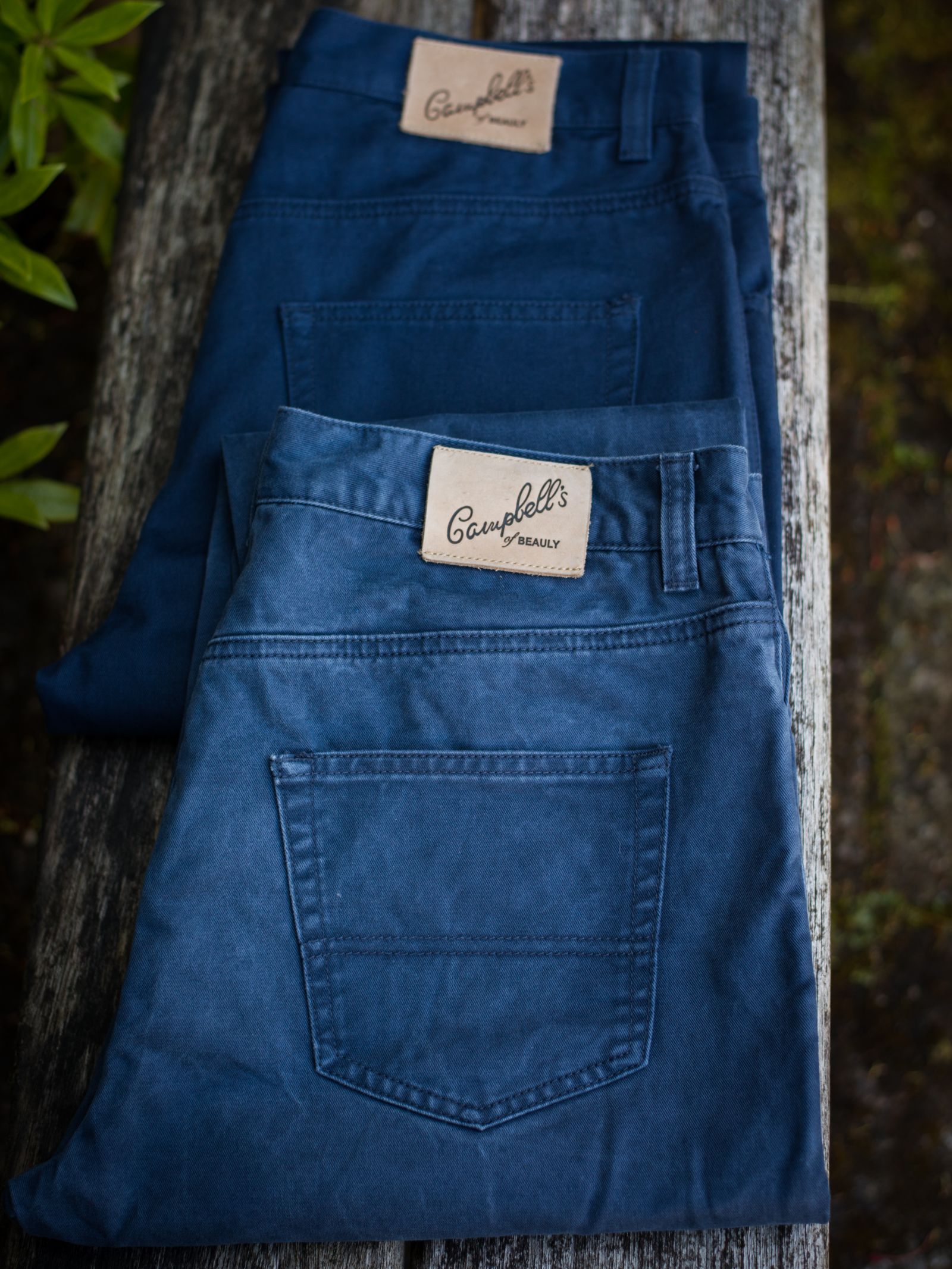 Two pairs of blue Colour Cotton Jeans by Campbell's of Beauly, each with a five-pocket design and tan leather patch on the waistband, are folded on a weathered wooden surface.