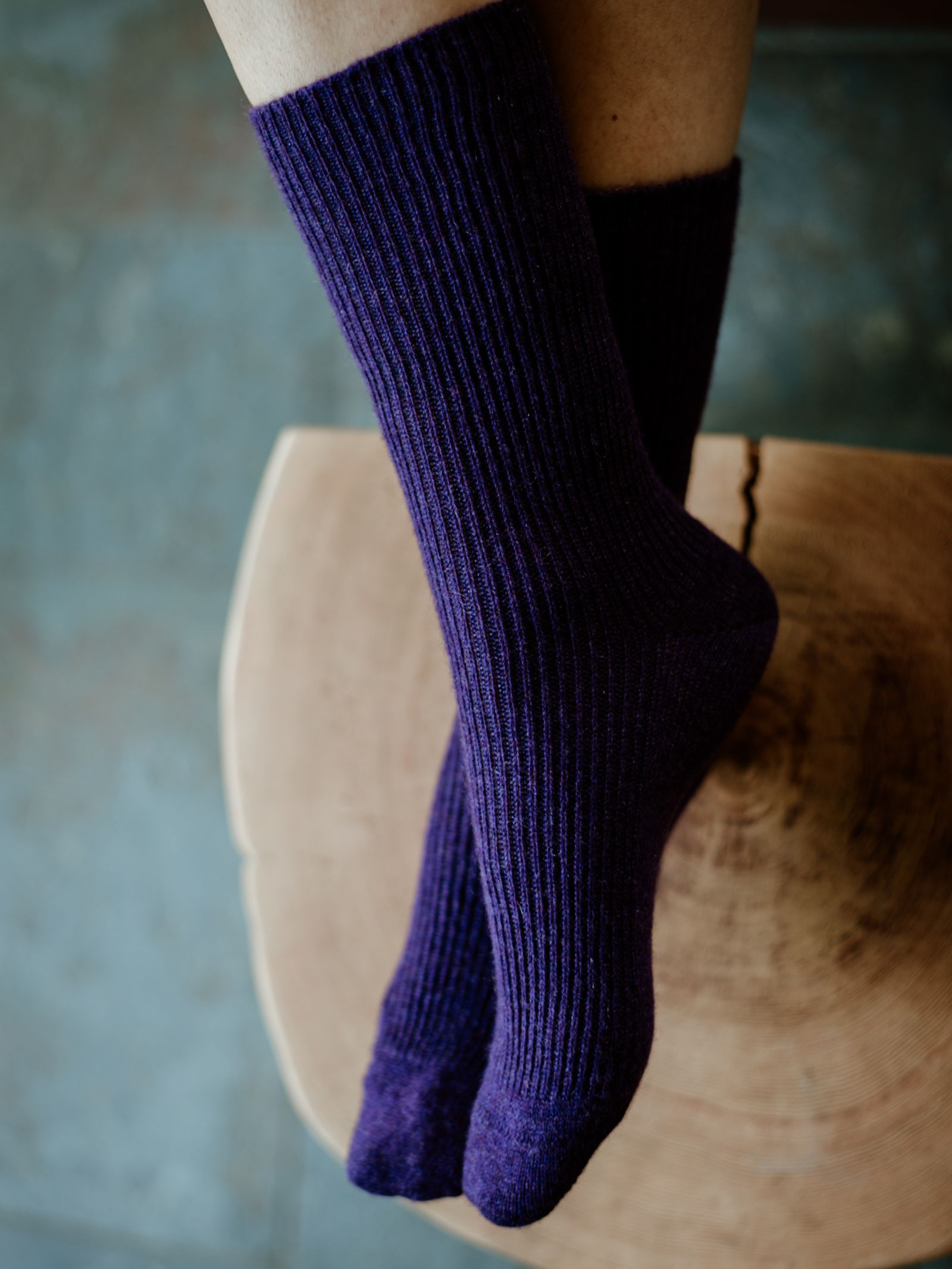 A person in thick, ribbed, dark purple Plain Cashmere Socks by Campbells of Beauly sits with crossed ankles on wood—a cozy luxury gift from the Scottish Border—set against a softly blurred background.