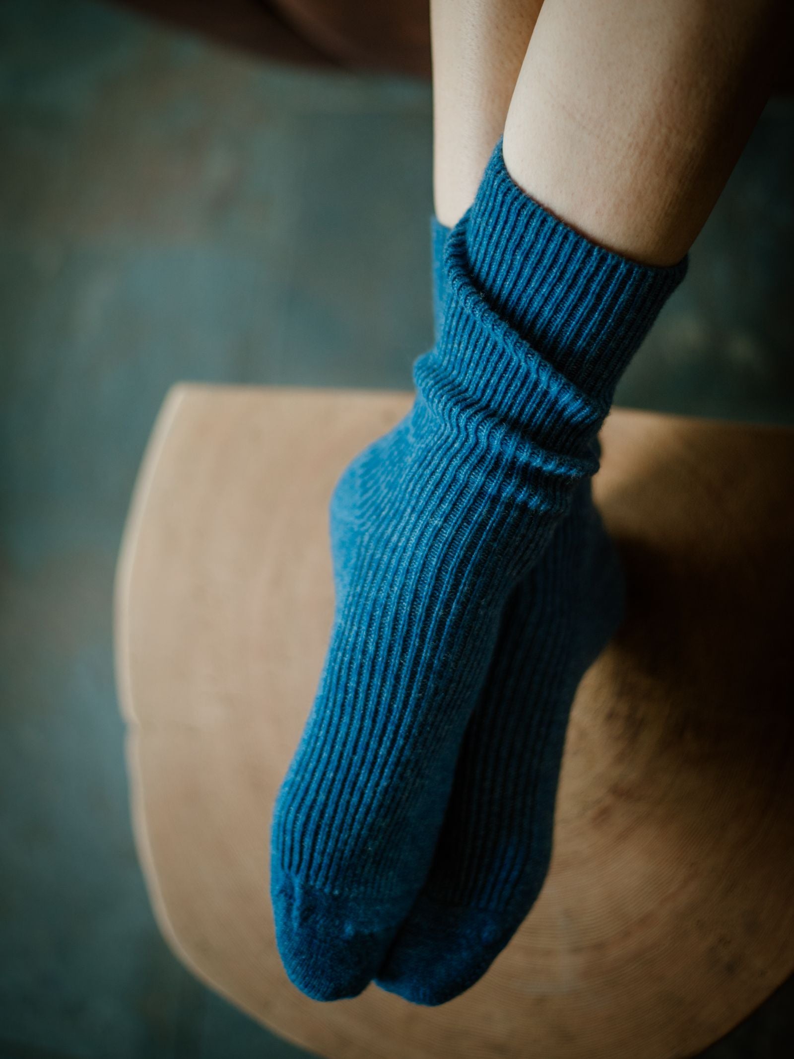 Someone wearing Campbells of Beauly Plain Cashmere Socks in blue relaxes with their feet on a wooden surface, legs partially visible and background softly blurred—capturing the luxurious comfort of these premium Scottish socks.