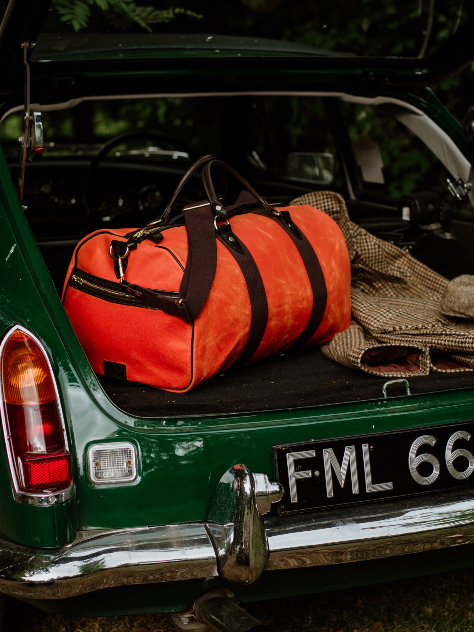 A Campbell's of Beauly Wax Holdall with bridle leather handles rests in the open trunk of a vintage green car, beside a checked blanket and hat. The license plate reads FML 66.