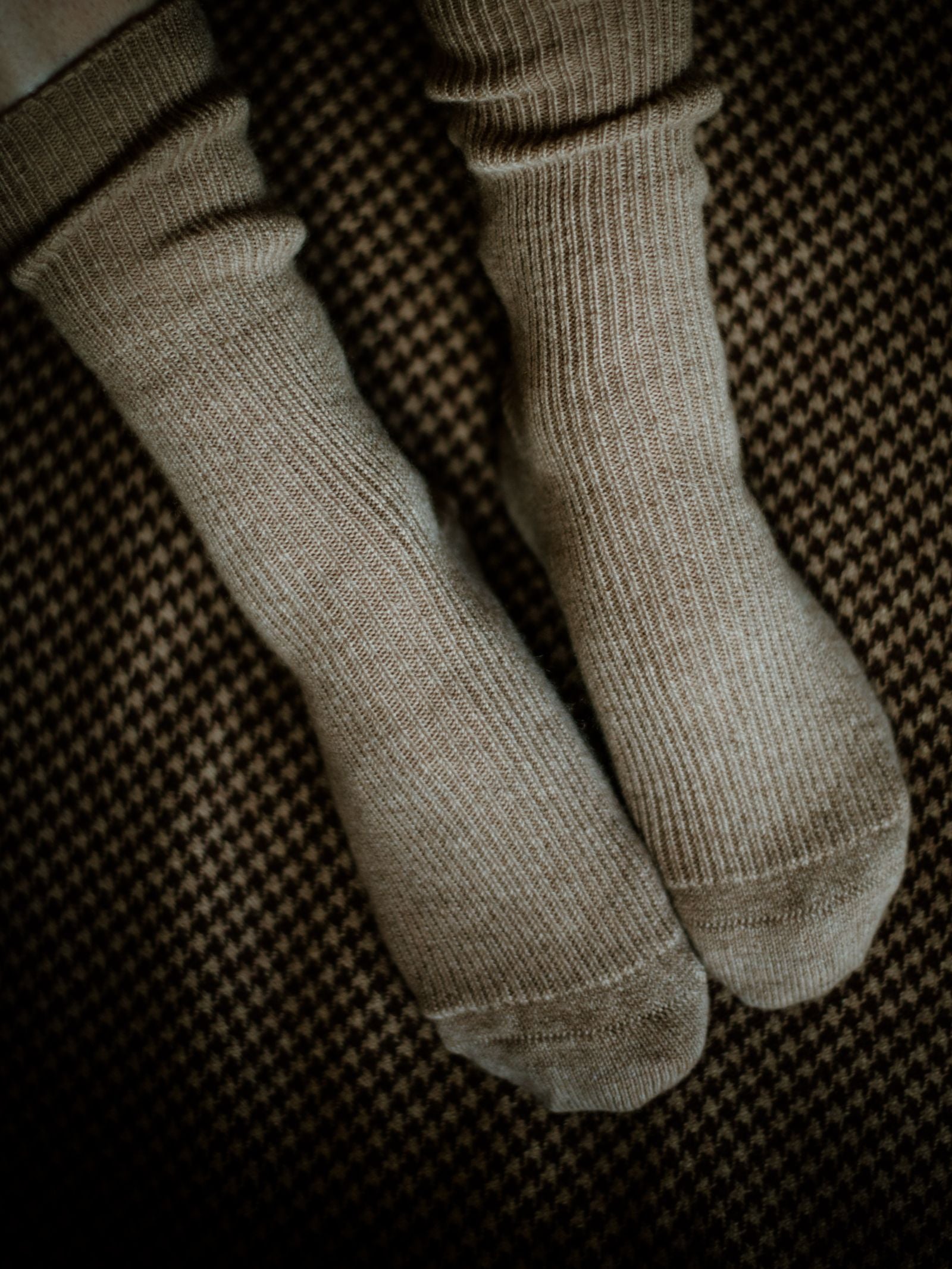 A close-up of two feet together in light brown Campbells of Beauly Plain Cashmere Socks on a dark, small-patterned carpet—softly lit for a cozy, luxurious gift from the Scottish Borders.