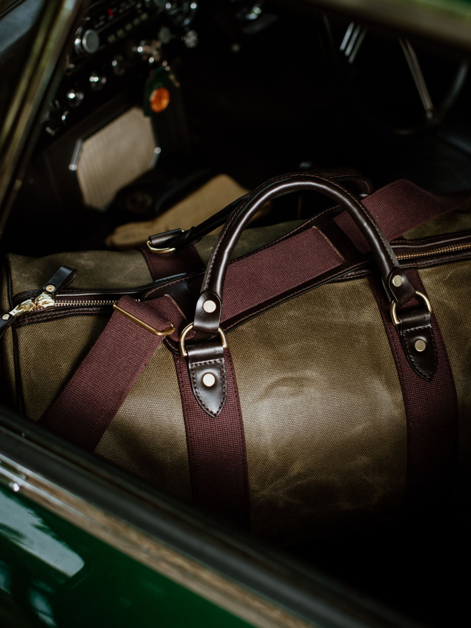 A close-up of the Campbell's of Beauly Wax Holdall—green waxed canvas with brown leather handles and maroon straps—rests on a classic car's passenger seat, dashboard and steering wheel in view: the perfect stylish travel companion.