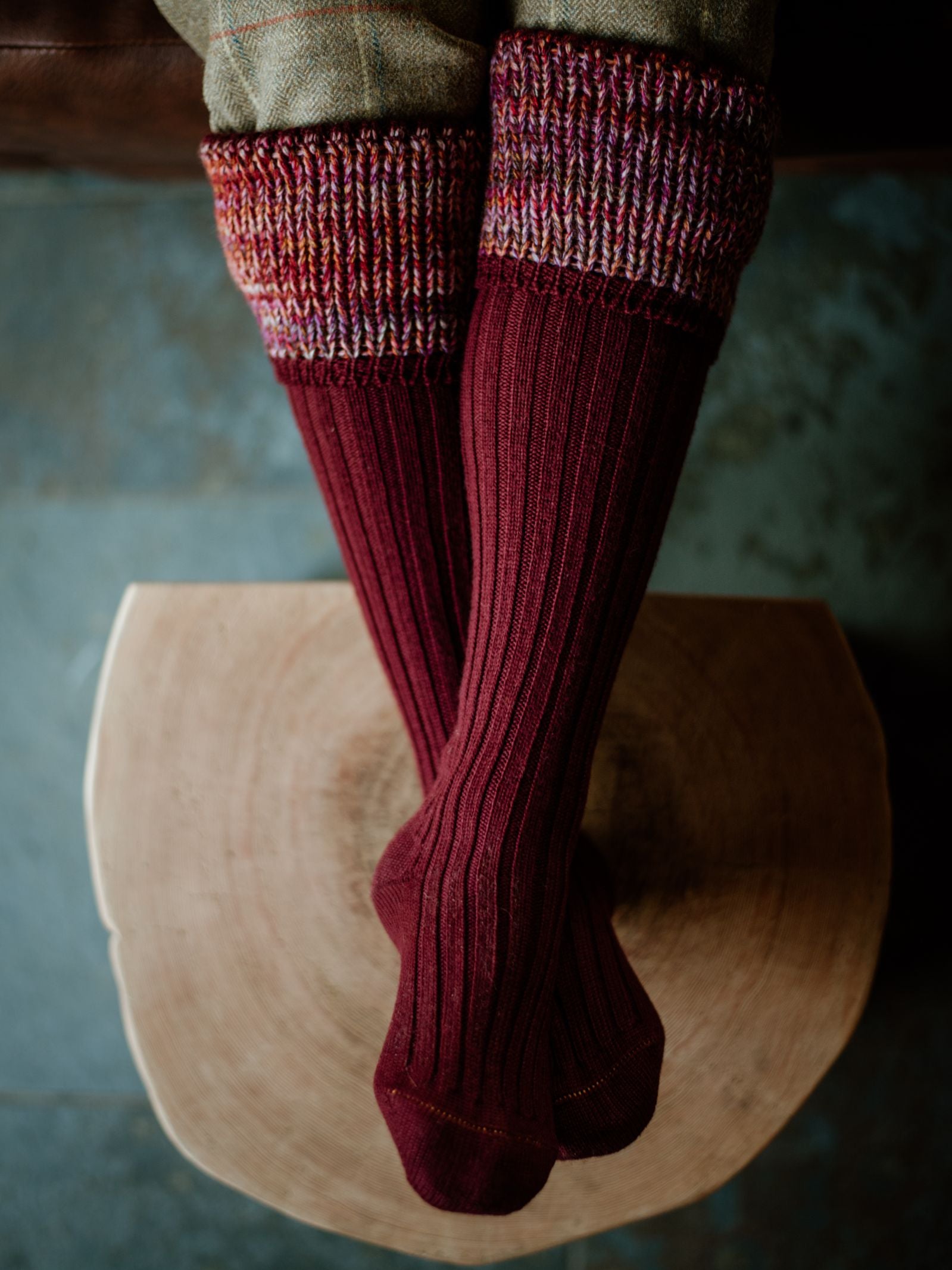 A person sits cross-legged on a wooden stool, seen from above against a blue-toned backdrop, wearing Campbells of Beauly Merino Katrine Shooting Socks with colorful cuffs—an elevated addition to any outdoor wardrobe.