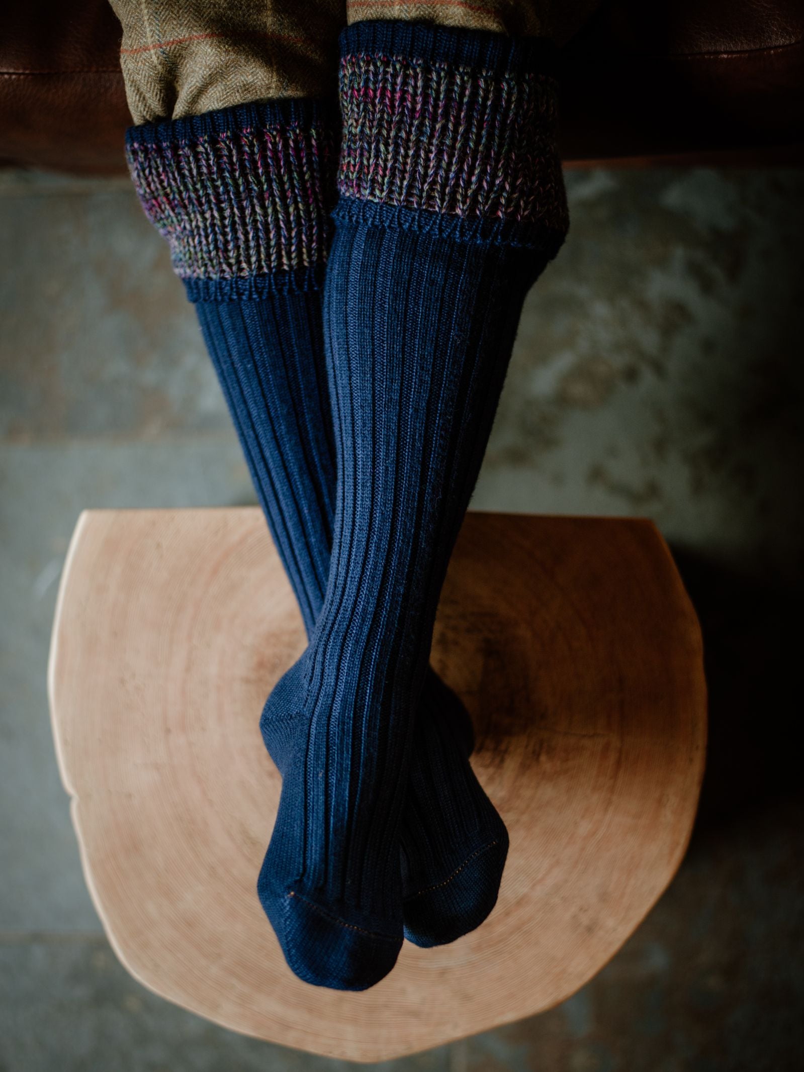 Someone wearing Campbells of Beauly Merino Katrine Shooting Socks in blue with multicolored cuffs rests their feet on a round wooden stool, seen from above.