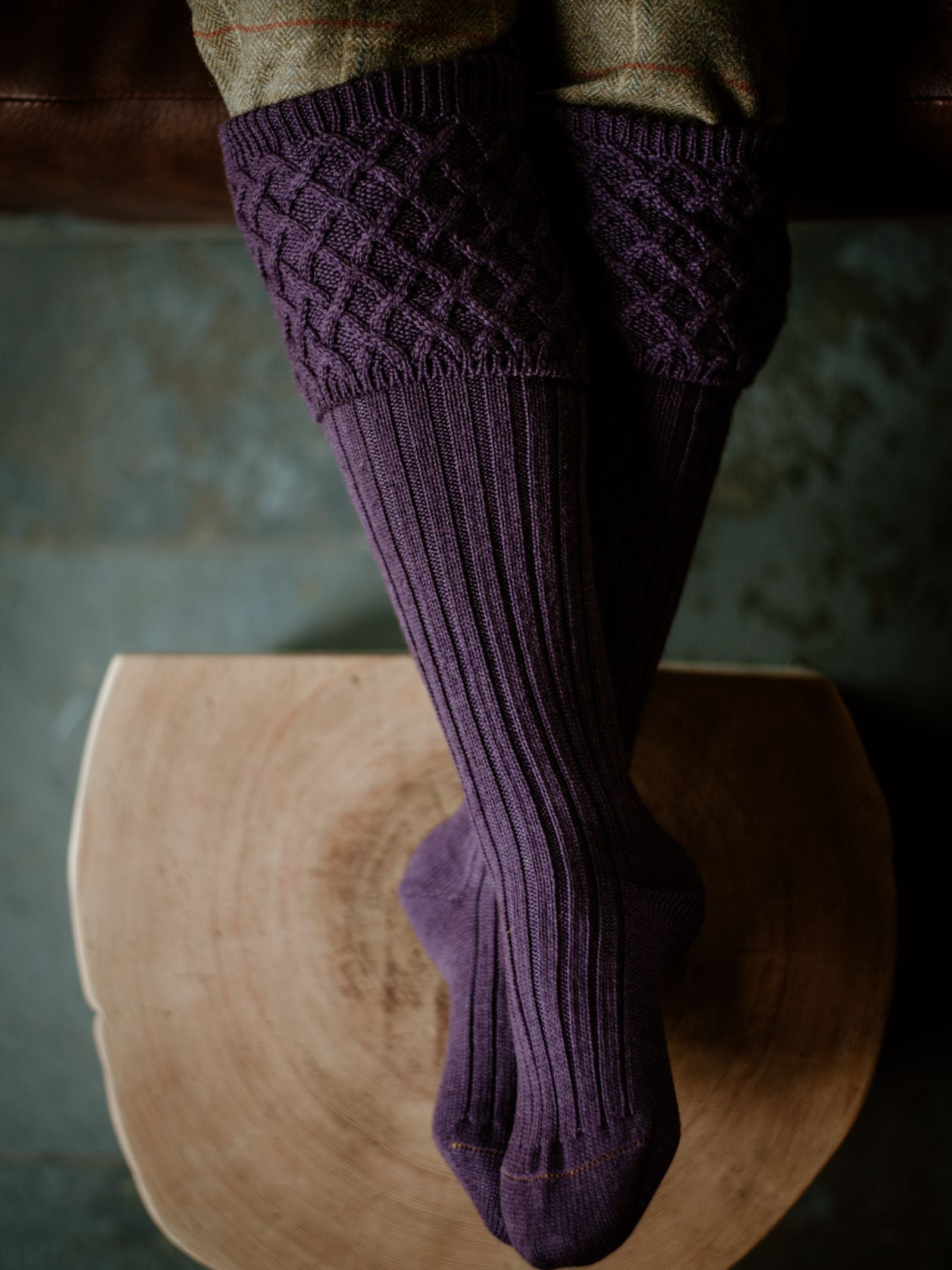 A person sits cross-legged on a wooden stool, wearing Campbells of Beauly Rannoch Merino Shooting Socks in purple with a textured diamond knit pattern, viewed from above.