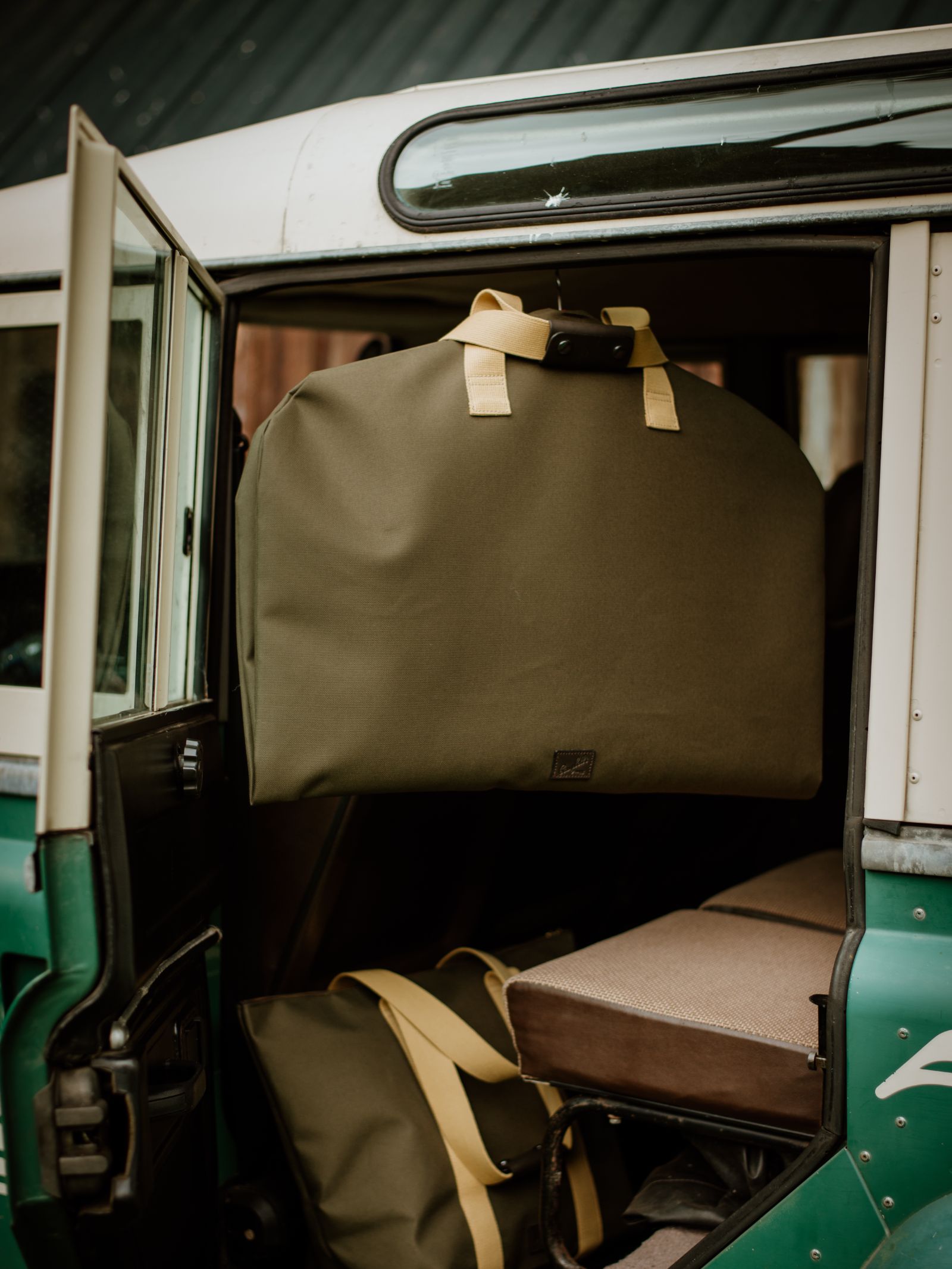 A Campbell's of Beauly Utility Suit Bag in olive green heavy-duty canvas with beige handles hangs from a vehicle door, while a matching garment-protecting bag rests on the seat inside.