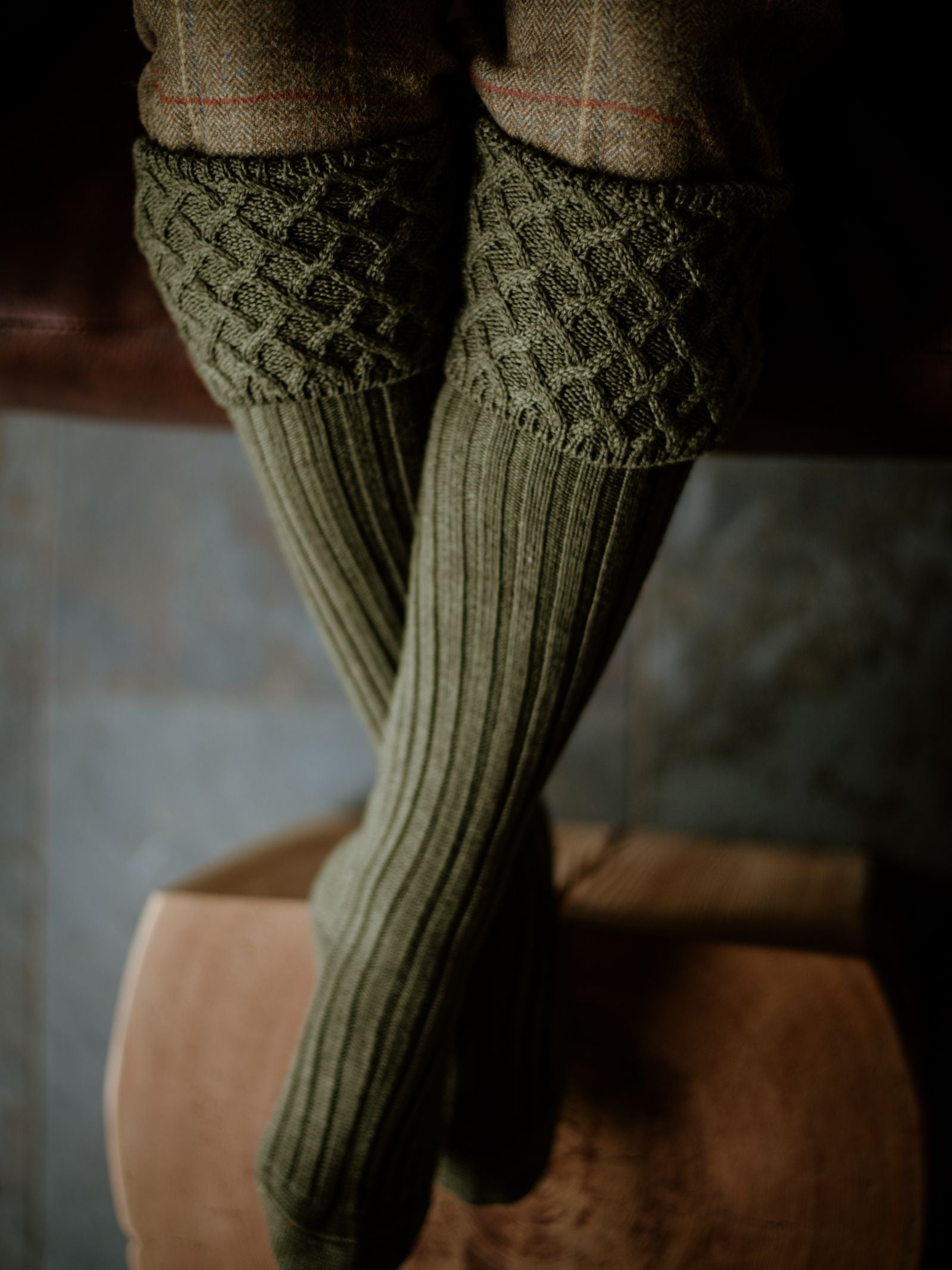 A close-up of a person seated with legs crossed, wearing Campbells of Beauly Rannoch Merino Shooting Socks in textured olive-green merino wool, paired with brown plaid trousers on a wooden surface against a blurred grey background.