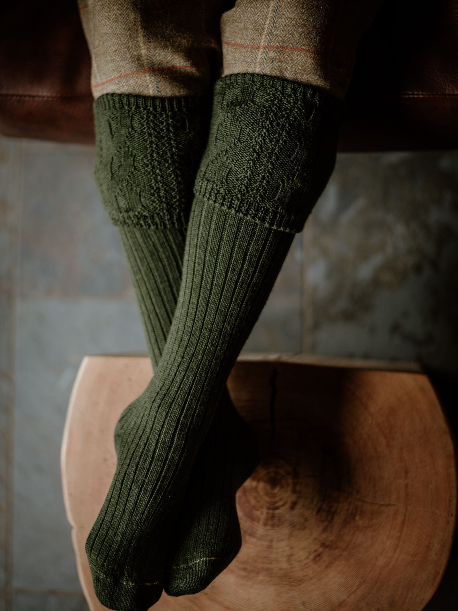 A person sits cross-legged on a brown wooden stool, wearing Campbells of Beauly Merino Glenmore Shooting Socks in green with a cable knit pattern. The look is completed with tweed trousers, channeling classic Scottish Borders style.