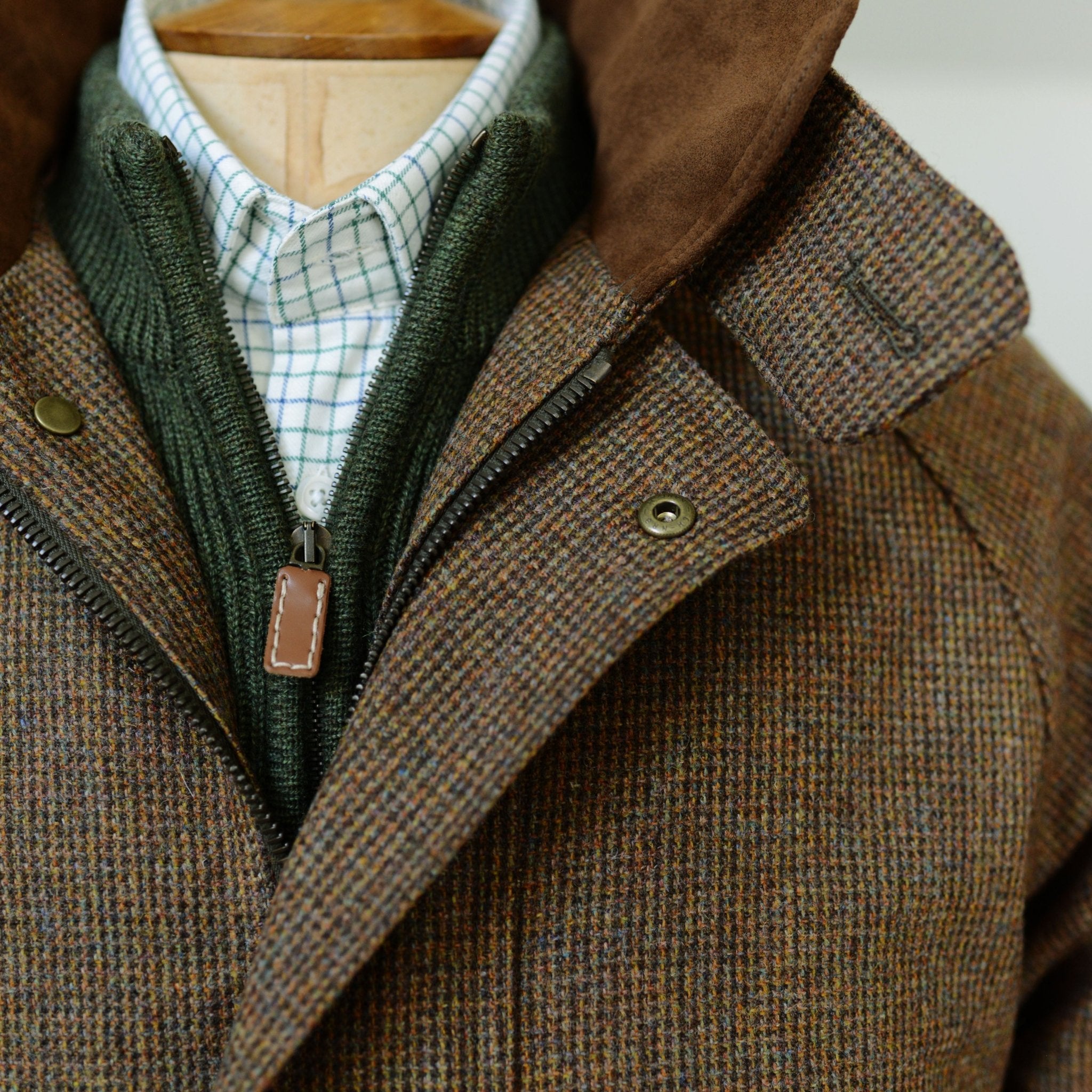A close-up of the Campbells of Beauly Tweed Field Coat with a brown plaid pattern, Alcantara collar, and metallic snaps, styled with a green zip-up sweater and white shirt featuring a blue check.