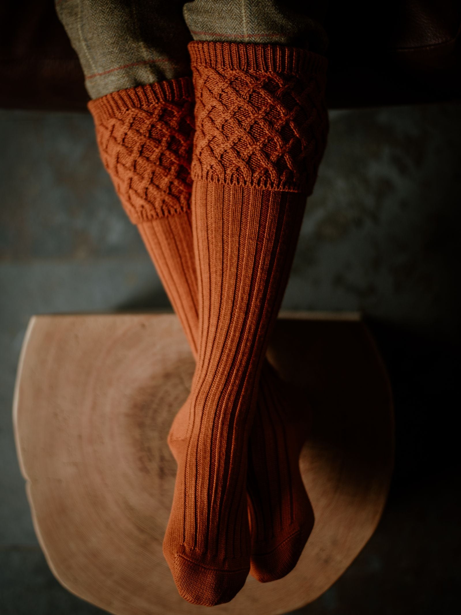 A person sits cross-legged on a wooden seat wearing Campbells of Beauly Rannoch Merino Shooting Socks in orange, featuring a textured diamond pattern at the top and ribbed legs.
