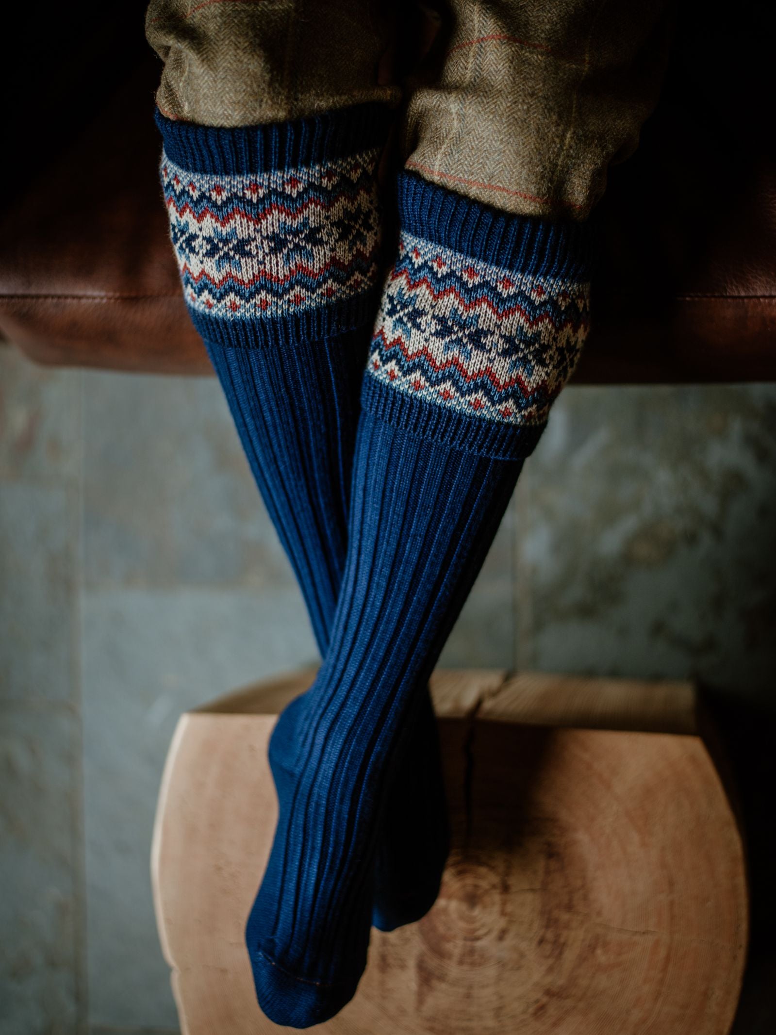 A person wearing Campbells of Beauly Merino Fairisle Shooting Socks, featuring a colorful patterned band, and brown pants sits cross-legged on a wooden stool.