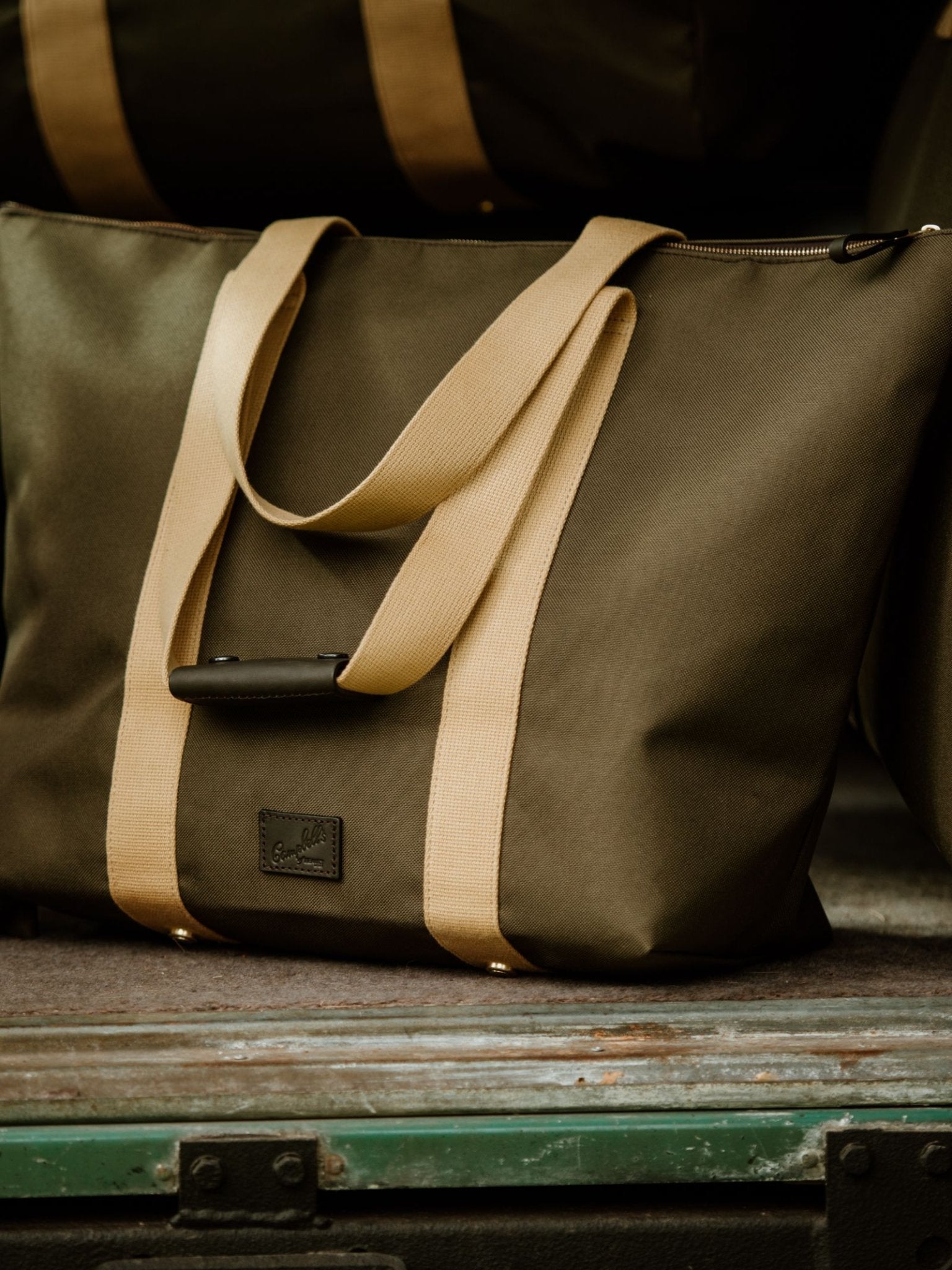 The Campbell's of Beauly Utility Medium Tote in olive green with beige straps and a black patch rests on a weathered green surface, making it an ideal everyday carryall, with other Utility Medium Totes visible in the background.