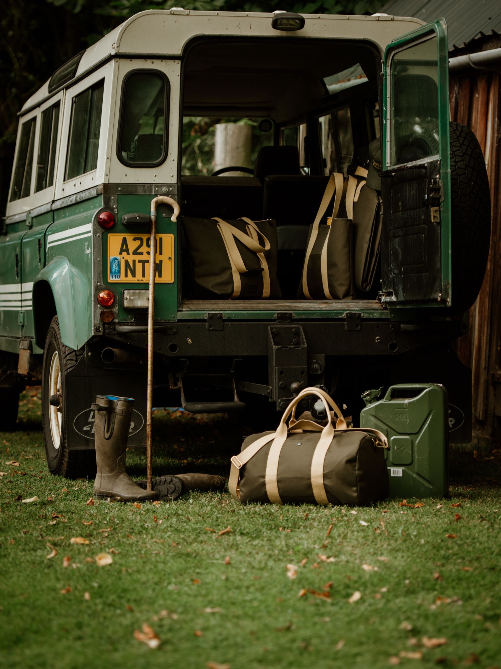A green Land Rover with open back doors is parked on grass. Behind it, a Campbell's of Beauly Utility Medium Duffle, a jerry can, a walking stick, and muddy boots are arranged. Trees and a wooden shed are in the background.