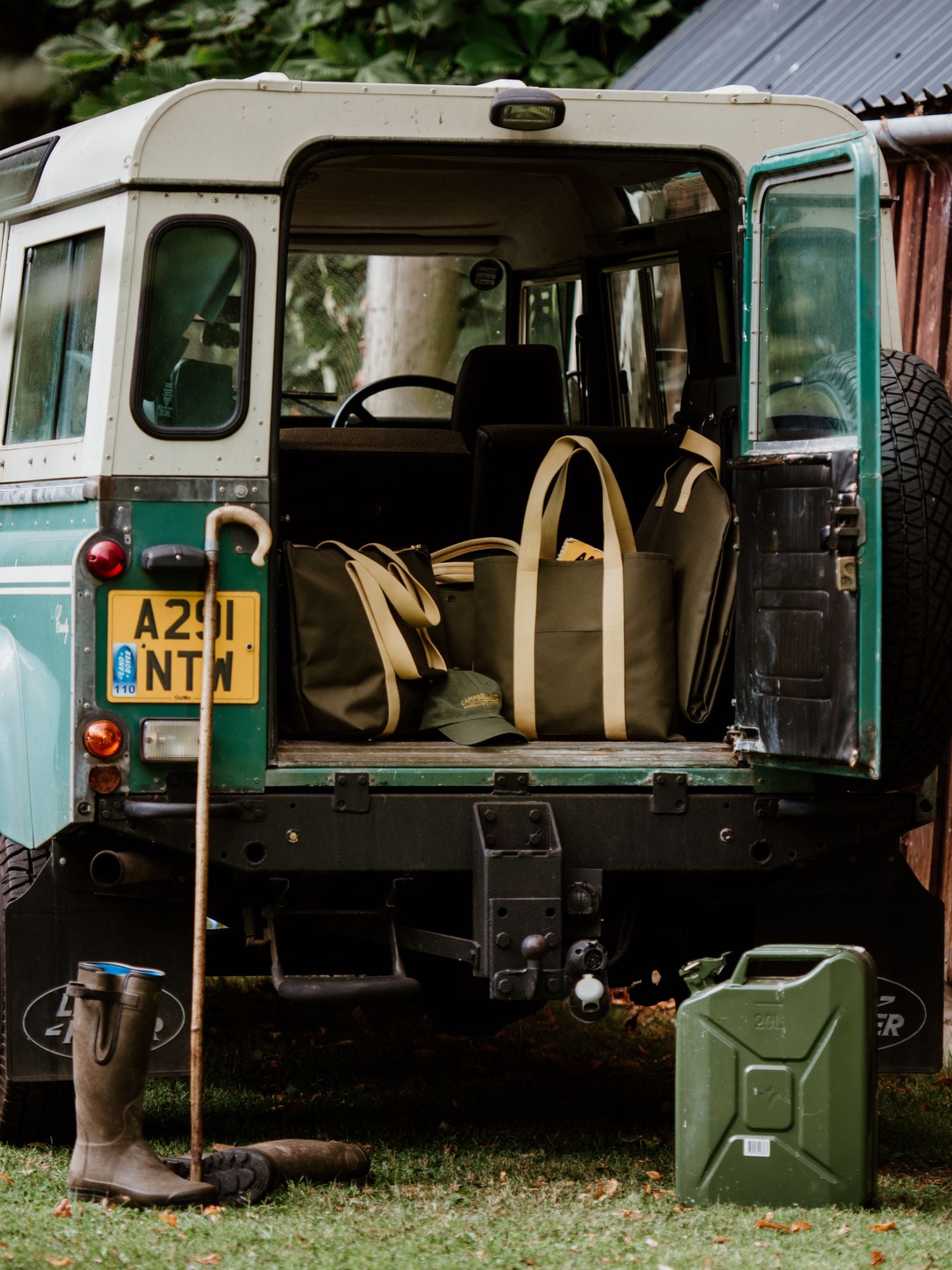 An open green and white Land Rover with canvas bags in the back. On the grass behind it are a walking stick, muddy boots, and the Campbell’s of Beauly Connoisseur Upcycled Mini Bar jerry can. Trees and a rustic building appear in the background.