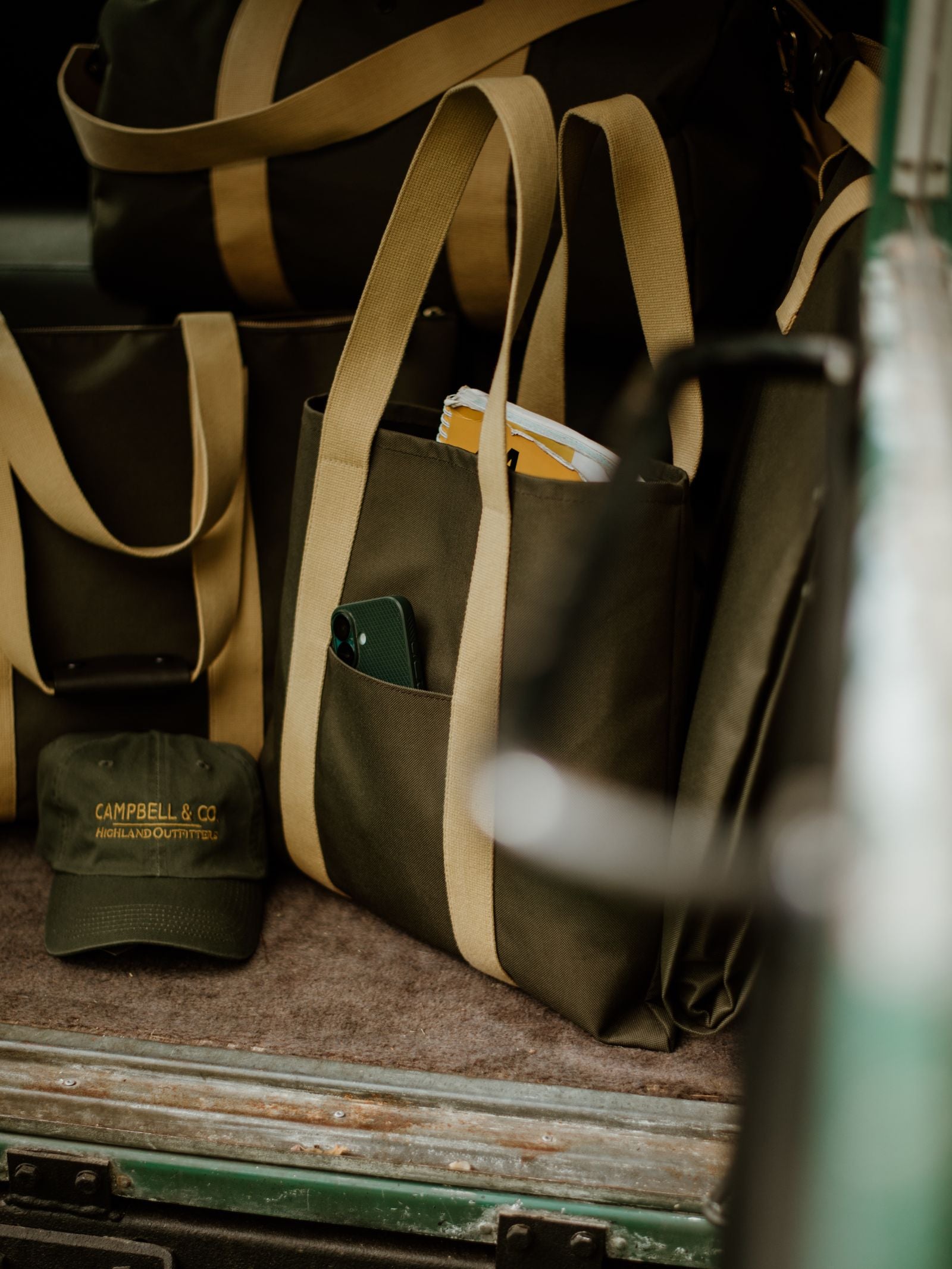 A close-up of Campbell's of Beauly Utility Packaway Tote bags in olive green with beige straps, one holding a phone and papers in a vehicle. An olive green “Campbell & Co” cap is seen on the floor. Ideal for everyday use.