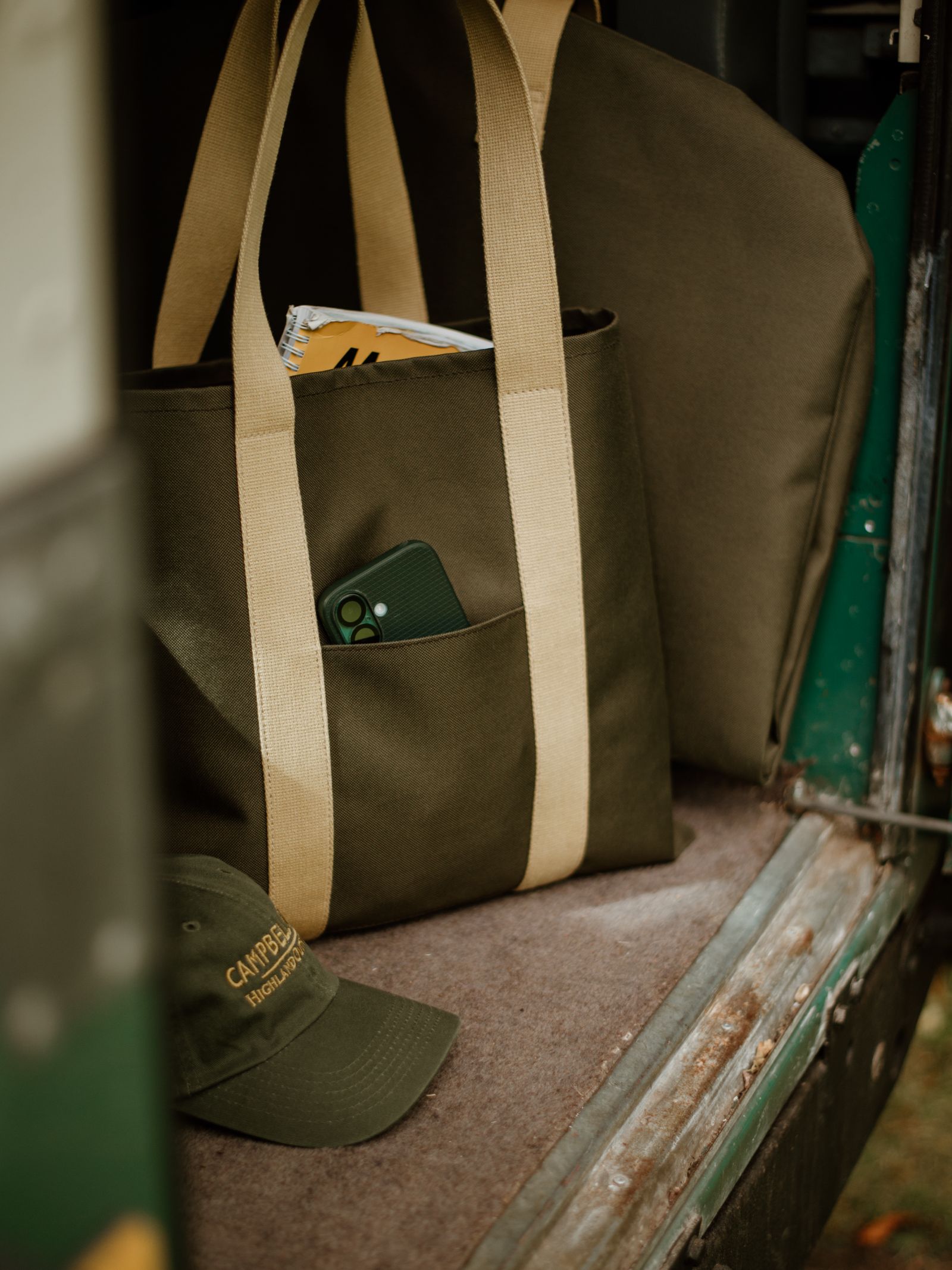 A green Campbell's of Beauly Utility Packaway Tote with beige straps rests in a vehicle, front pocket holding a green smartphone and a folded yellow map inside. Nearby, a green cap completes this perfect everyday utility bag setup.