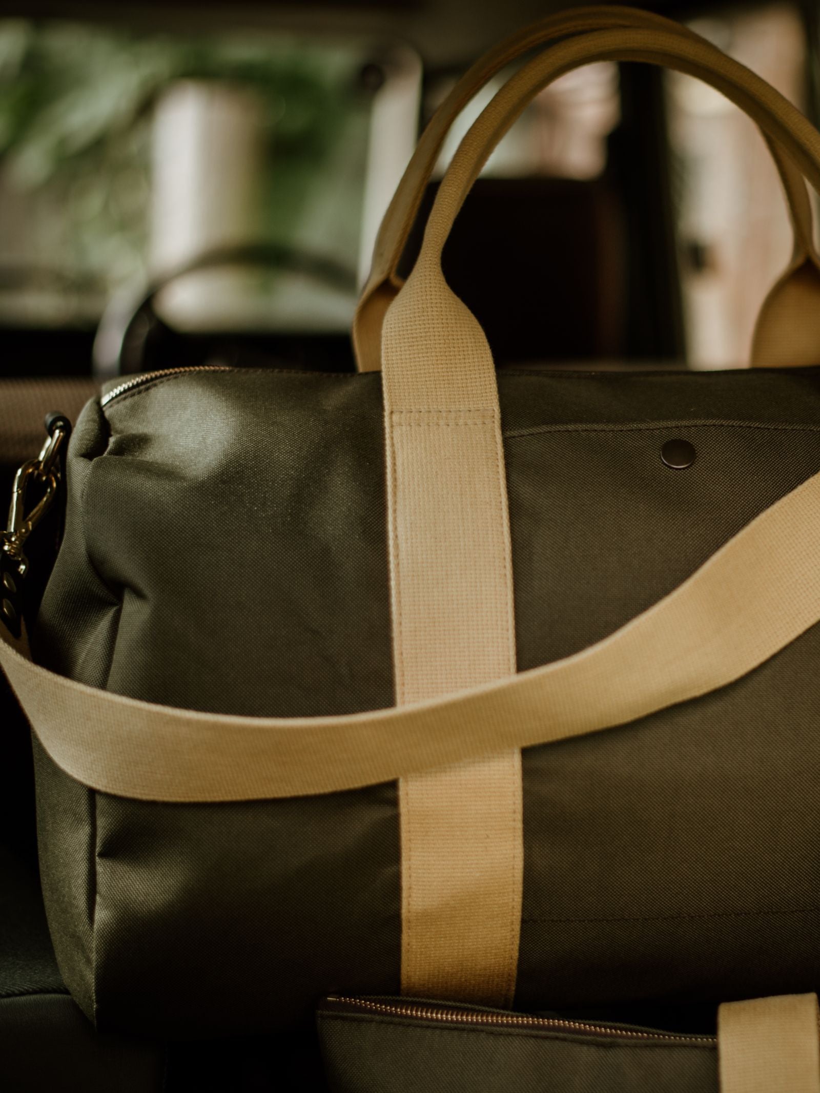 The Campbell's of Beauly Utility Medium Duffle in olive green canvas with tan straps sits on a car seat against a blurred background, evoking a sense of travel or adventure.
