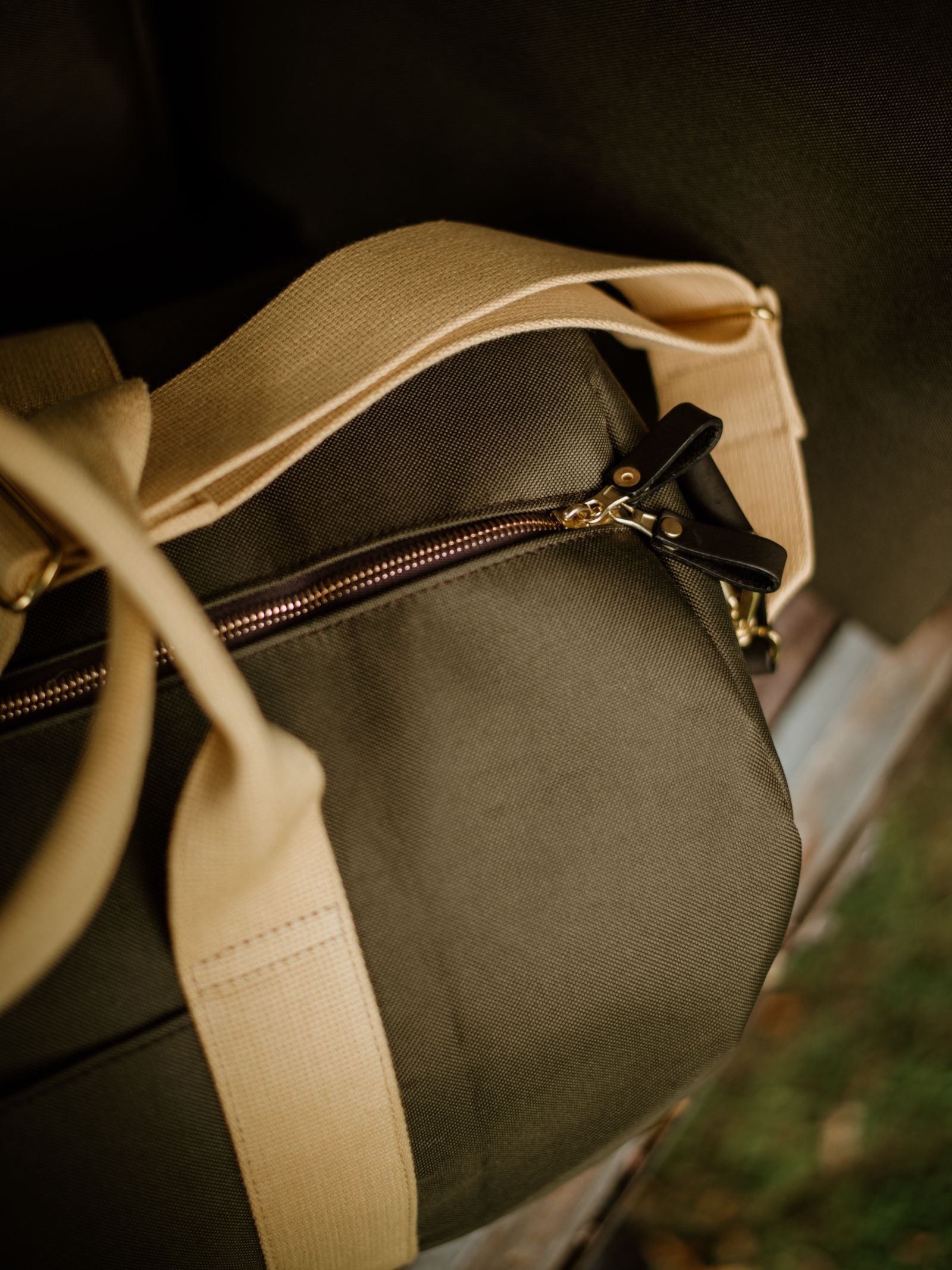 A close-up of the Campbell's of Beauly Utility Medium Duffle in olive green canvas with beige straps and a gold zipper sits partially unzipped on a wooden surface, with grass visible below.
