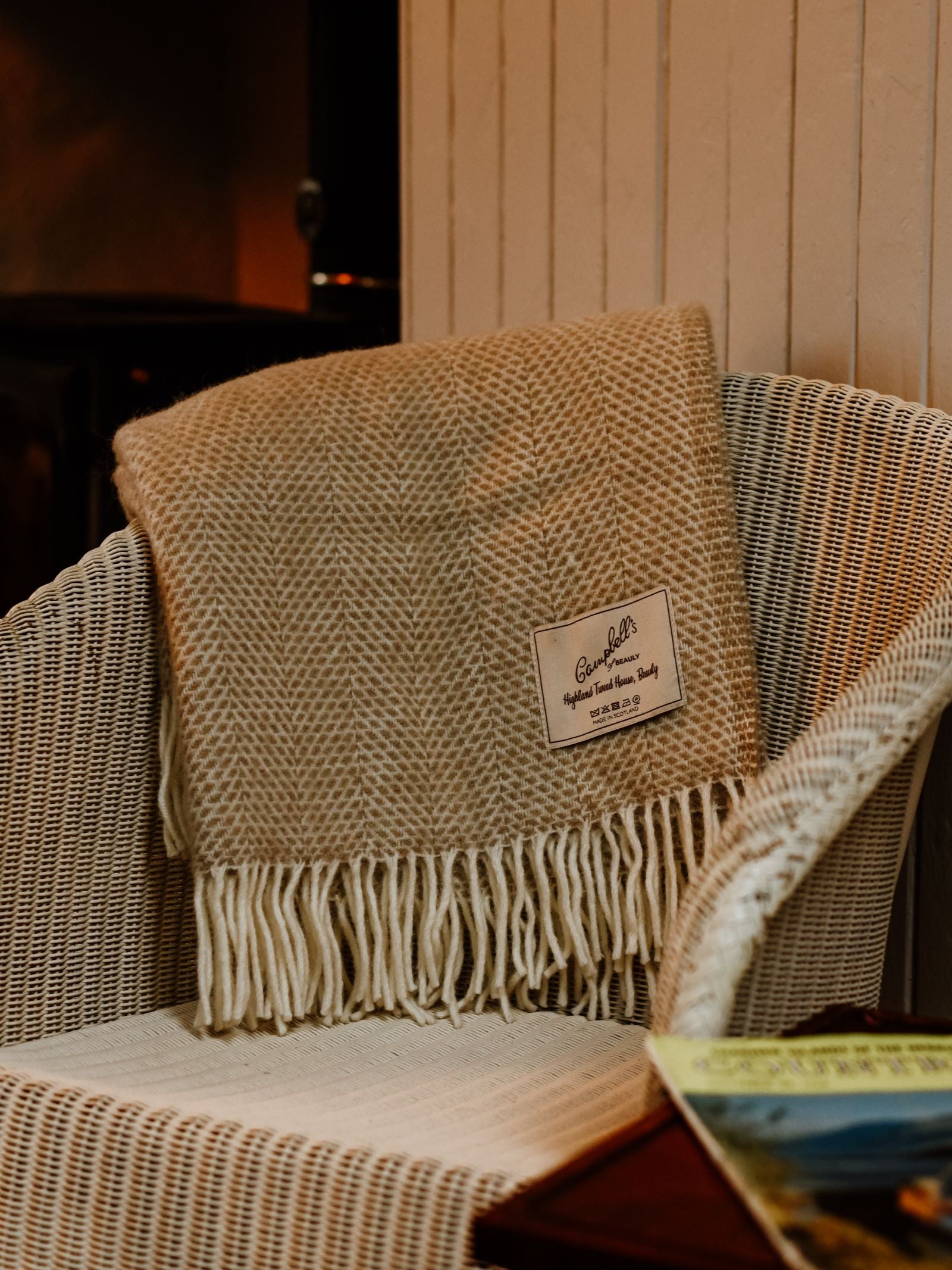 The Stipled Twill Blanket by Campbell's of Beauly, in beige with fringe and stitched label, drapes over a white wicker chair. Its breathable twill weave offers comfort and style, with a magazine resting on a wooden table nearby.