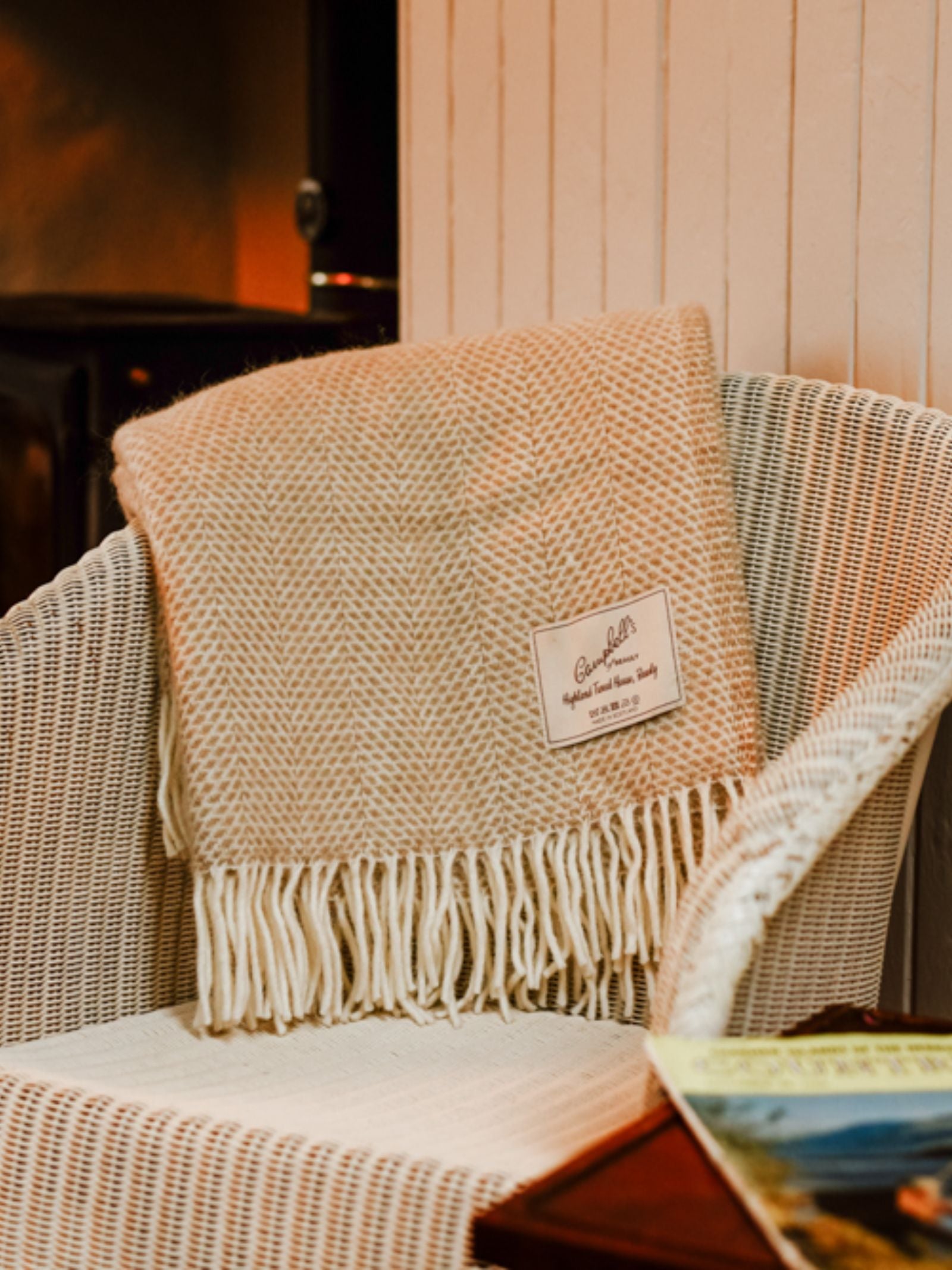 A cream Stipled Twill Blanket by Campbell's of Beauly with fringe and stitched label drapes over a white wicker chair. Part of a magazine sits on the adjacent wooden table.